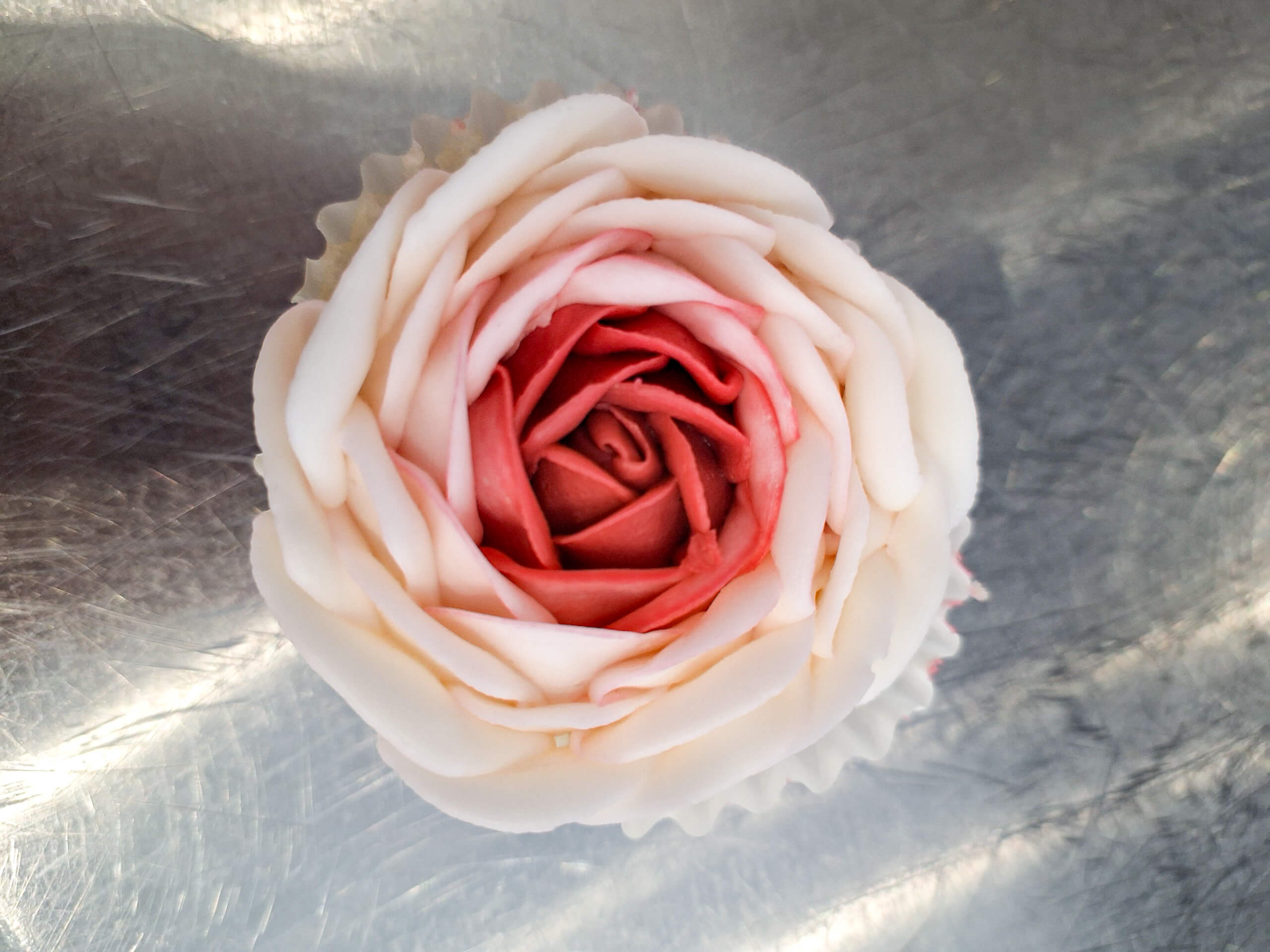 Dutch Heritage Bakery Sturgis MI A cupcake topped with pink and white frosting arranged to resemble a rose, placed on a metallic surface.