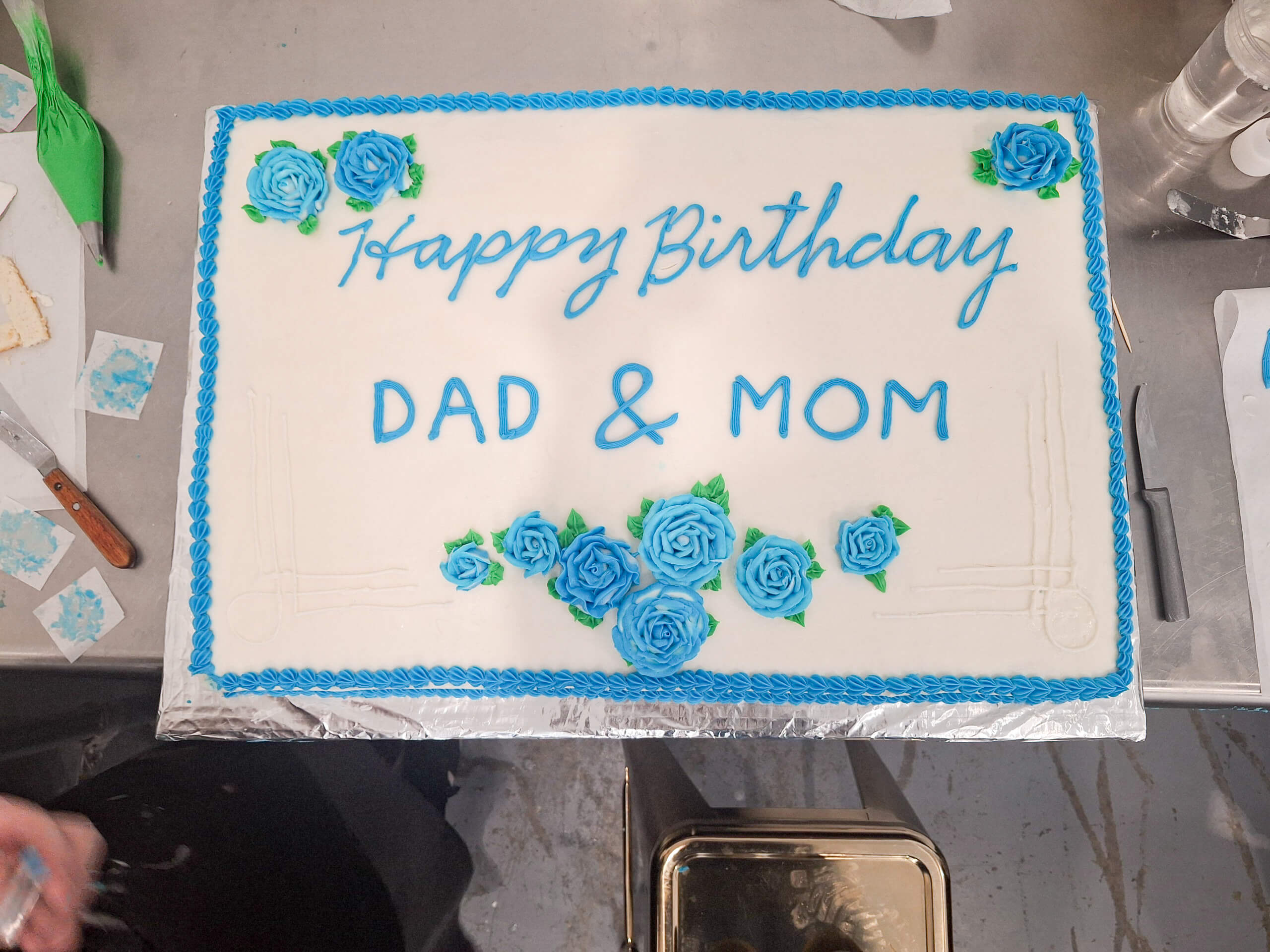 Dutch Heritage Bakery Sturgis MI A rectangular birthday cake with white icing, blue piped writing that says "Happy Birthday DAD & MOM," and decorated with blue rose designs.