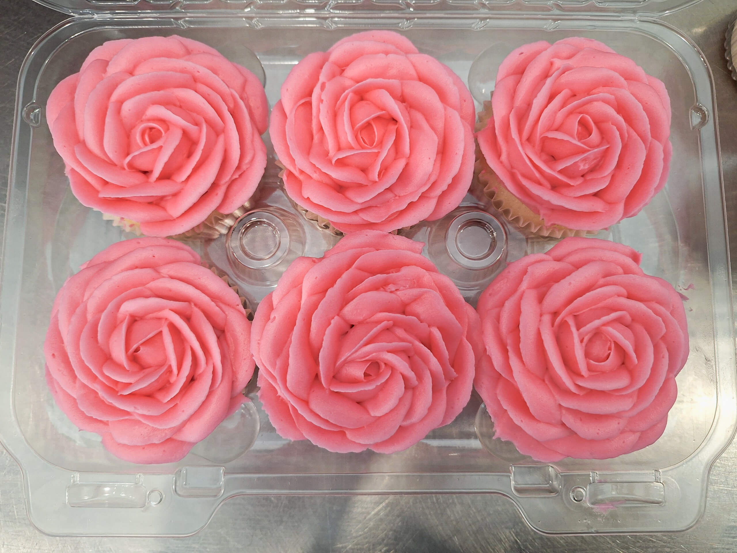 Dutch Heritage Bakery Sturgis MI Six cupcakes topped with pink rose-shaped frosting are arranged in a clear plastic container on a metallic surface.