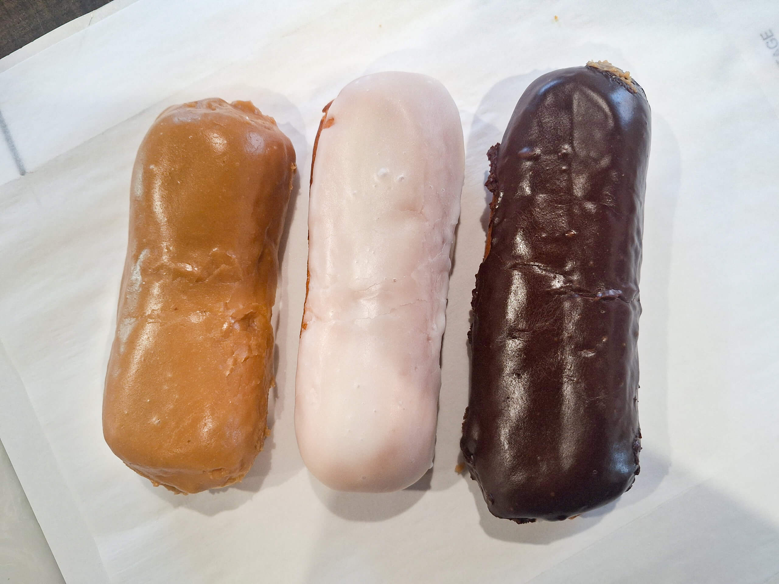 Dutch Heritage Bakery Sturgis MI Three long, rectangular donuts with different glazes—caramel, white, and chocolate—are arranged side by side on a white surface.