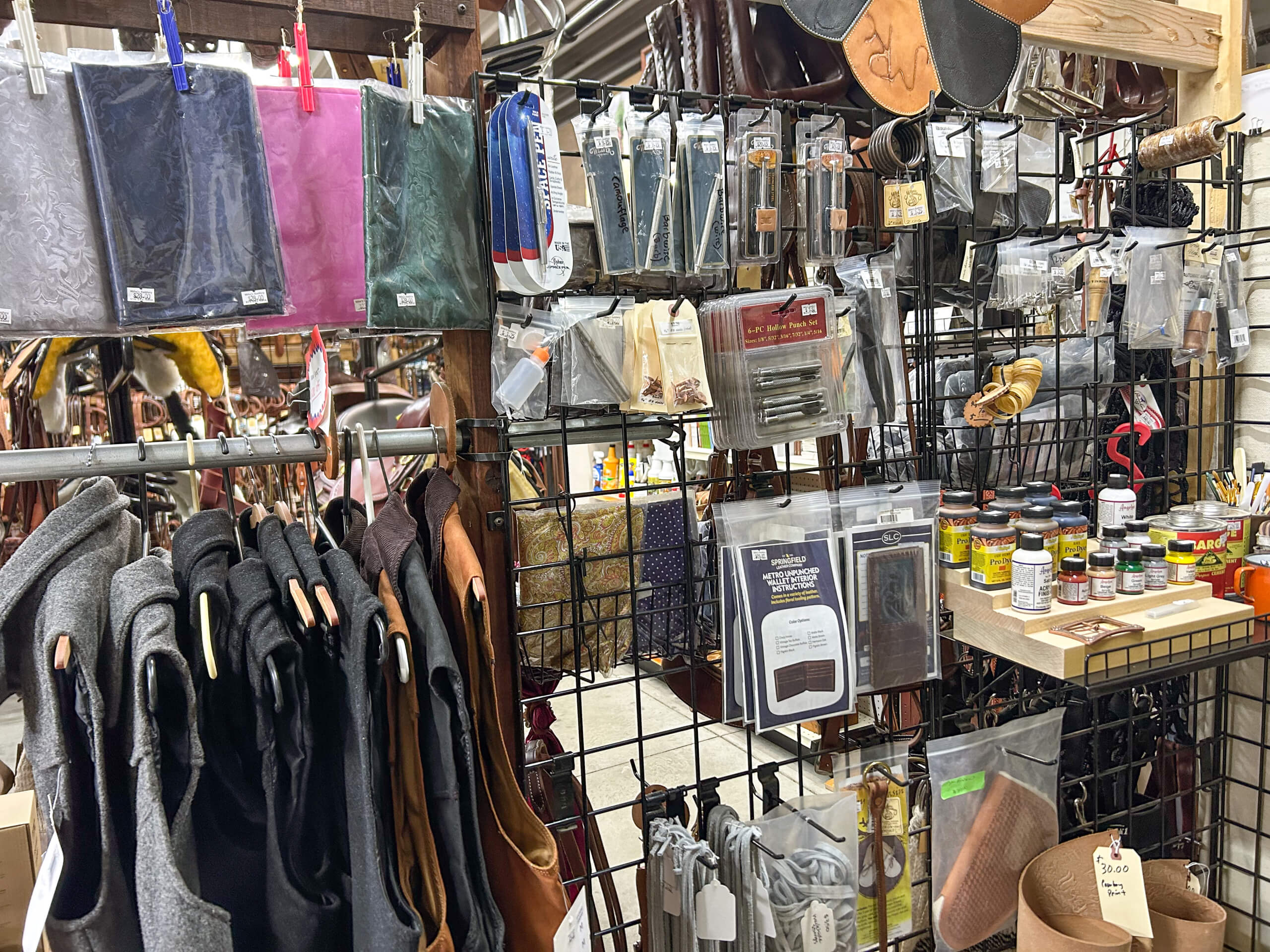 Miller's Saddlery Goshen IN A display of leatherworking supplies, including leather pieces, tools, dyes, wallets, and accessories, organized on black wire racks in a store.