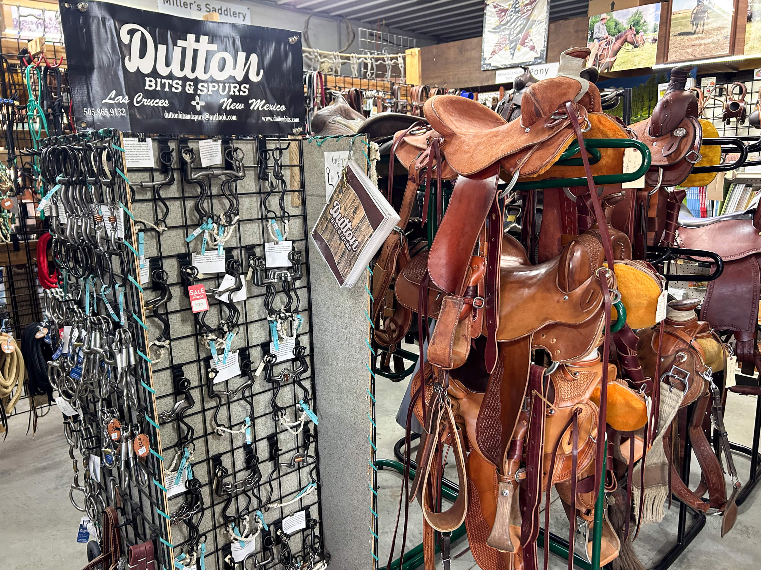 Miller's Saddlery Goshen IN A display of horse saddles, bits, and spurs inside a tack shop. Saddles hang on racks, while bits and spurs are arranged on a stand labeled "Dutton Bits & Spurs.