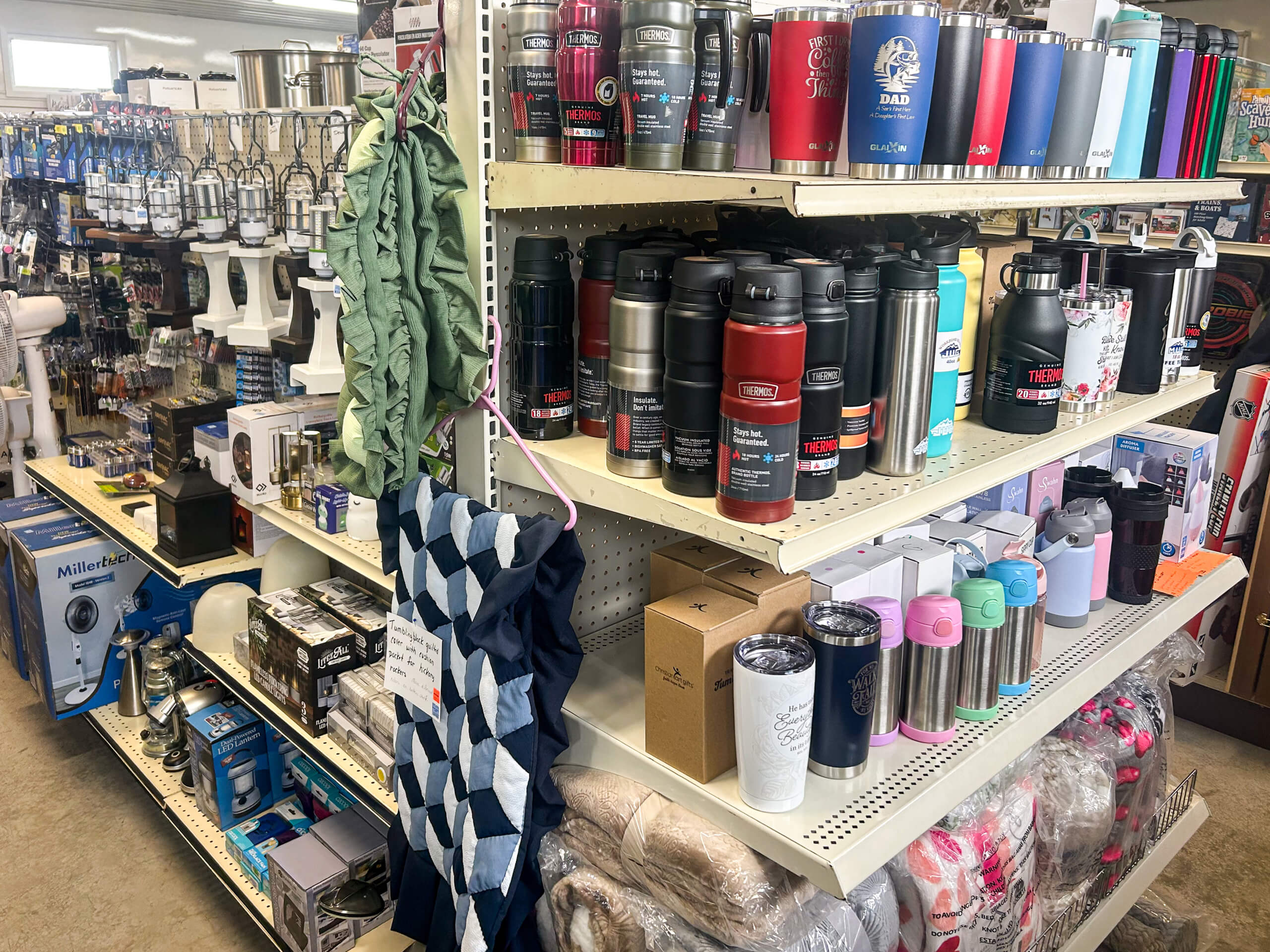 Miller's Variety Store Etna Green, IN Store aisle displaying insulated water bottles, thermoses, and tumblers on shelves, with various small appliances and household items visible in the background.