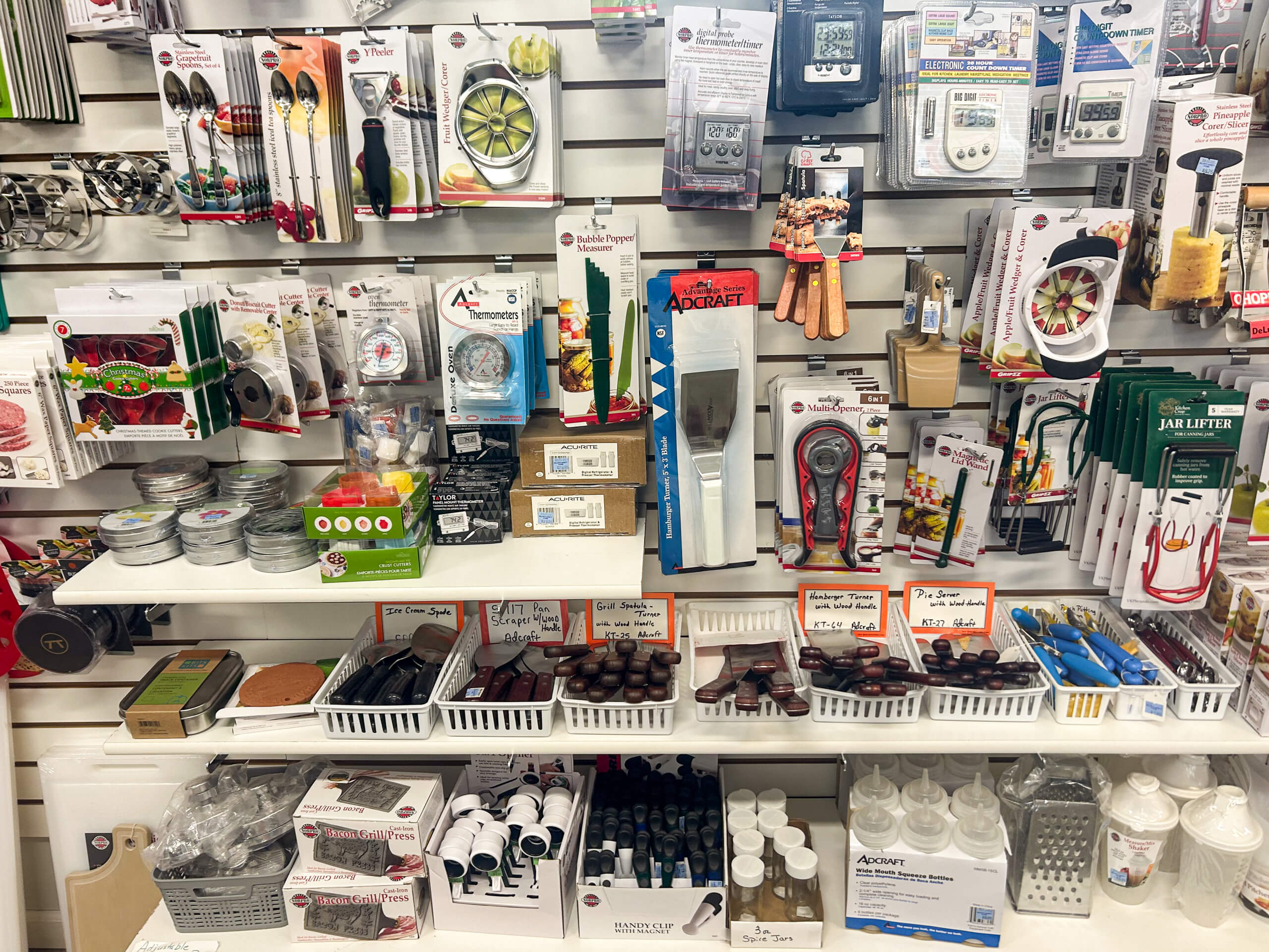 Miller's Variety Store Etna Green, IN A variety of kitchen gadgets and tools displayed on shelves and hanging on a white slat wall in a store. Items include timers, slicers, bottle openers, and small containers.