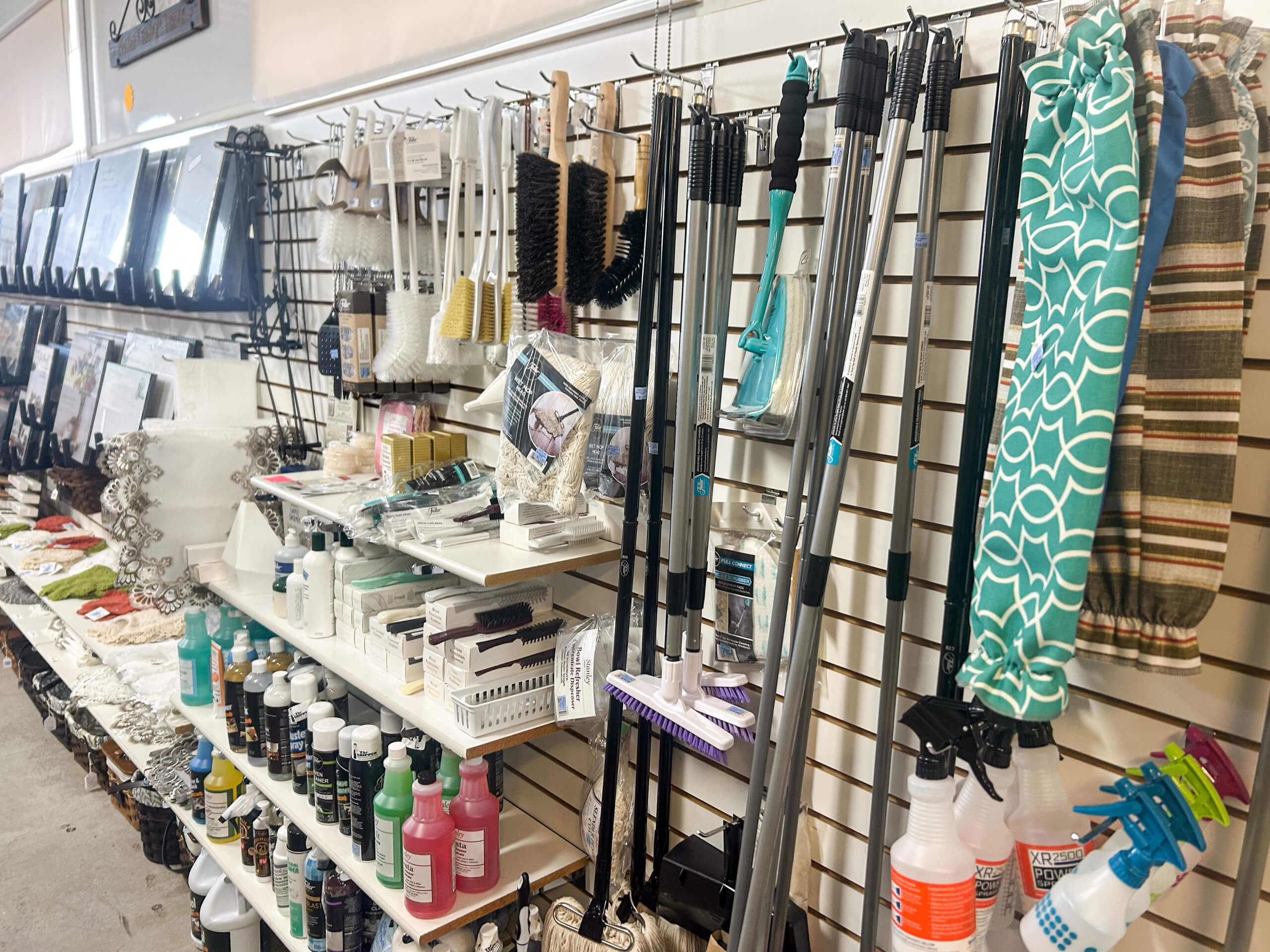 Miller's Variety Store Etna Green, IN A store aisle displays cleaning supplies, brushes, mops, spray bottles, laundry bags, and various household items organized on shelves and racks.