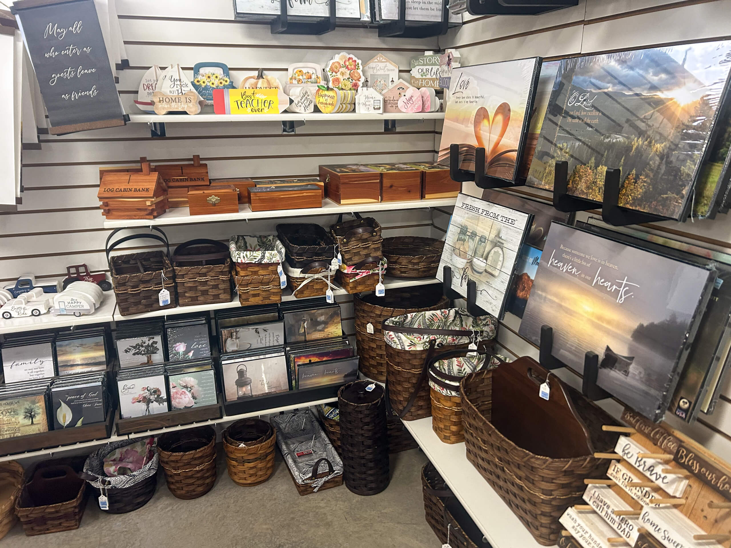 Miller's Variety Store Etna Green, IN Shelves display various home decor items including framed art, wooden boxes, woven baskets, signs with sayings, and greeting cards in a retail store setting.