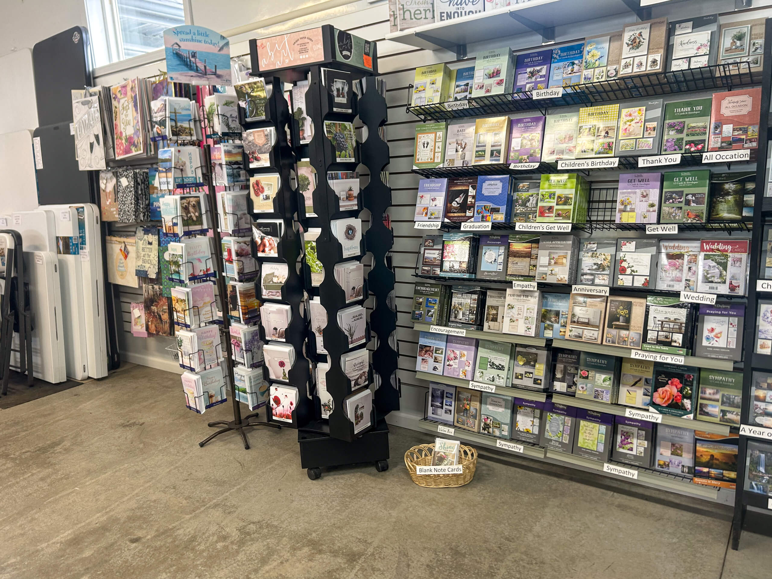Miller's Variety Store Etna Green, IN A display of greeting cards and gifts in a store, with shelves holding cards for various occasions and a rotating card stand in the center. A small basket is on the floor.