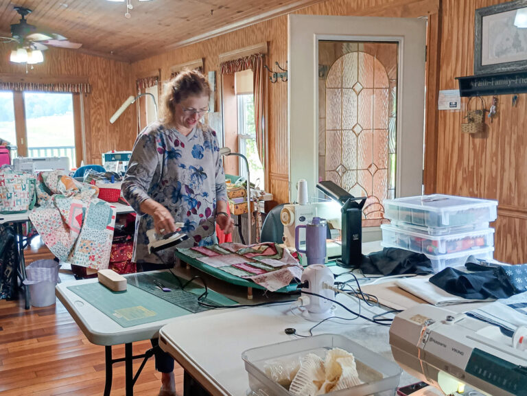 Weaver-View Farms Quilting Retreats Penn Yan, NY A woman irons a quilt piece in a craft room filled with sewing supplies, fabrics, and a sewing machine on tables.