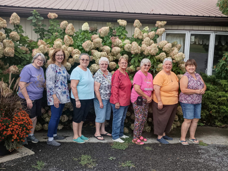 Weaver-View Farms Quilting Retreats Penn Yan, NY Eight women stand in a line outside in front of blooming hydrangea bushes and a beige building with a metal roof.