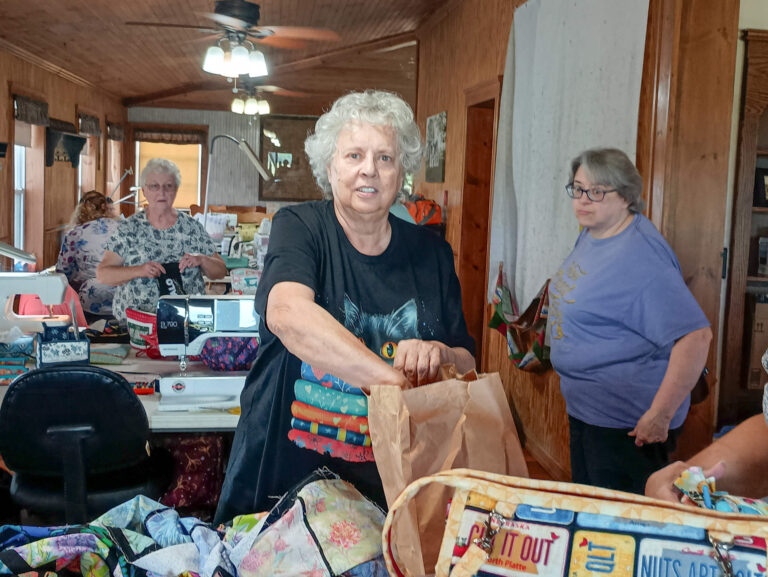 Weaver-View Farms Quilting Retreats Penn Yan, NY Three older women are in a room filled with quilting supplies; one woman stands at the center, packing a paper bag, while the others work or watch nearby.