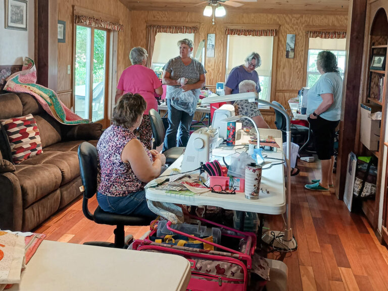 Weaver-View Farms Quilting Retreats Penn Yan, NY A group of women work on sewing projects around tables in a well-lit room with sewing machines, fabric, and supplies visible.