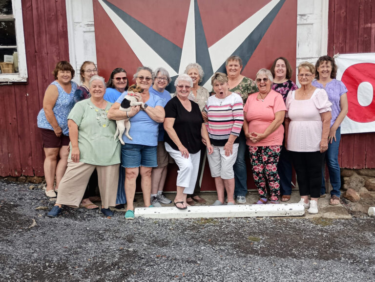 Weaver-View Farms Quilting Retreats Penn Yan, NY A group of thirteen women and one small dog pose and smile in front of a red barn with a large geometric design painted on the door.