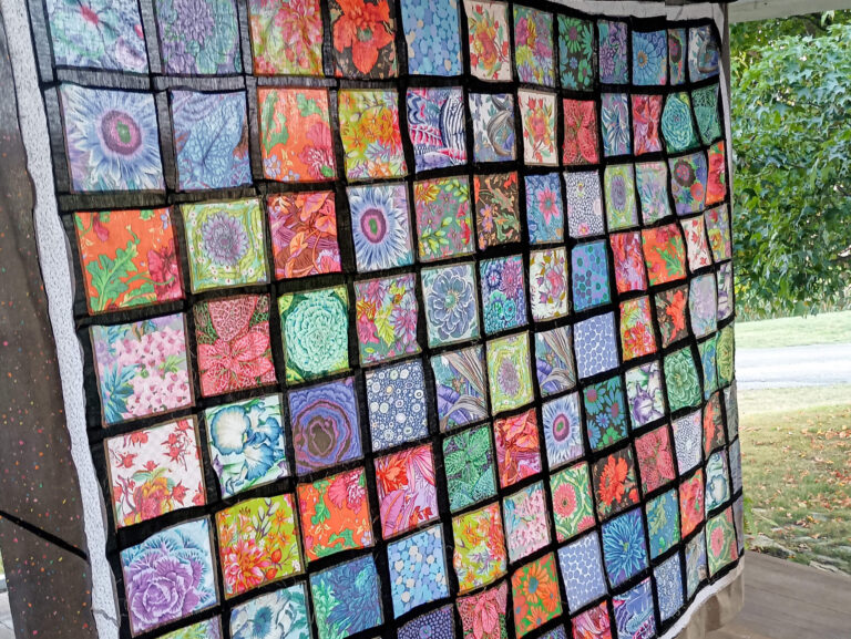 Weaver-View Farms Quilting Retreats Penn Yan, NY A colorful patchwork quilt with square panels featuring various floral and abstract designs, bordered by black lines, displayed outdoors near green trees.