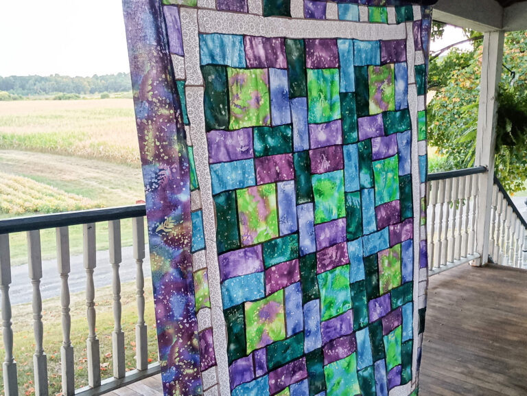 Weaver-View Farms Quilting Retreats Penn Yan, NY A colorful patchwork quilt with blue, green, purple, and black patterns hangs on a porch railing overlooking a rural field.