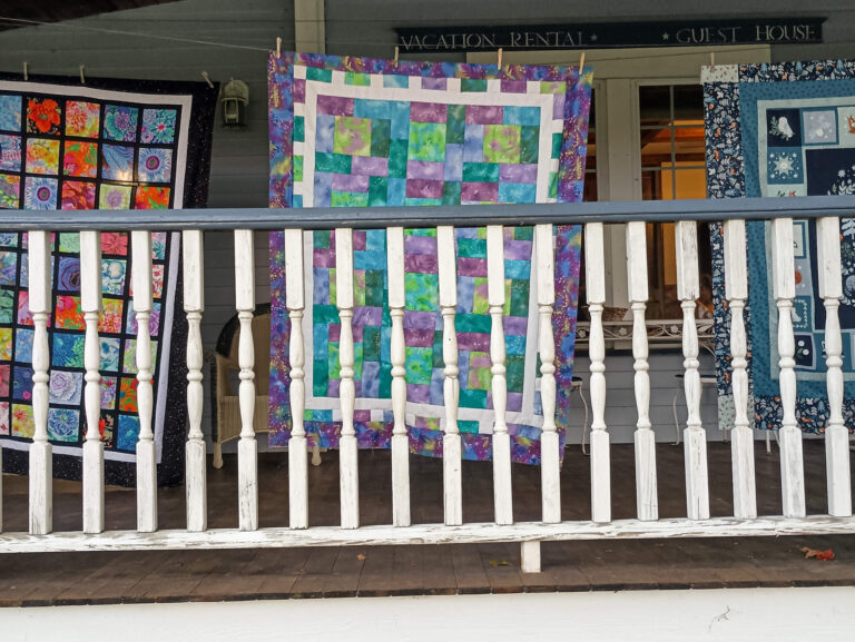 Weaver-View Farms Quilting Retreats Penn Yan, NY Three colorful patchwork quilts hang on the railing of a wooden porch in front of a building marked as a vacation rental and guest house.