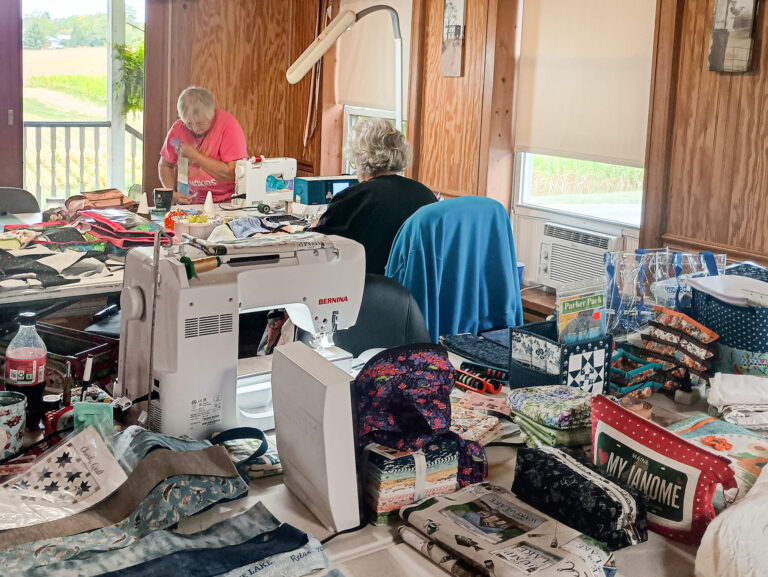 Weaver-View Farms Quilting Retreats Penn Yan, NY Two people work on sewing projects at a table covered with fabric, sewing machines, and quilting supplies in a sunlit room with wooden walls and windows.