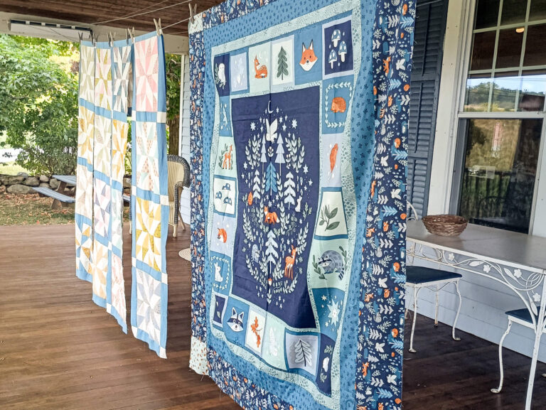 Weaver-View Farms Quilting Retreats Penn Yan, NY Three colorful patchwork quilts hang on a line on a wooden porch next to a white metal table and chair, with trees and a house window in the background.
