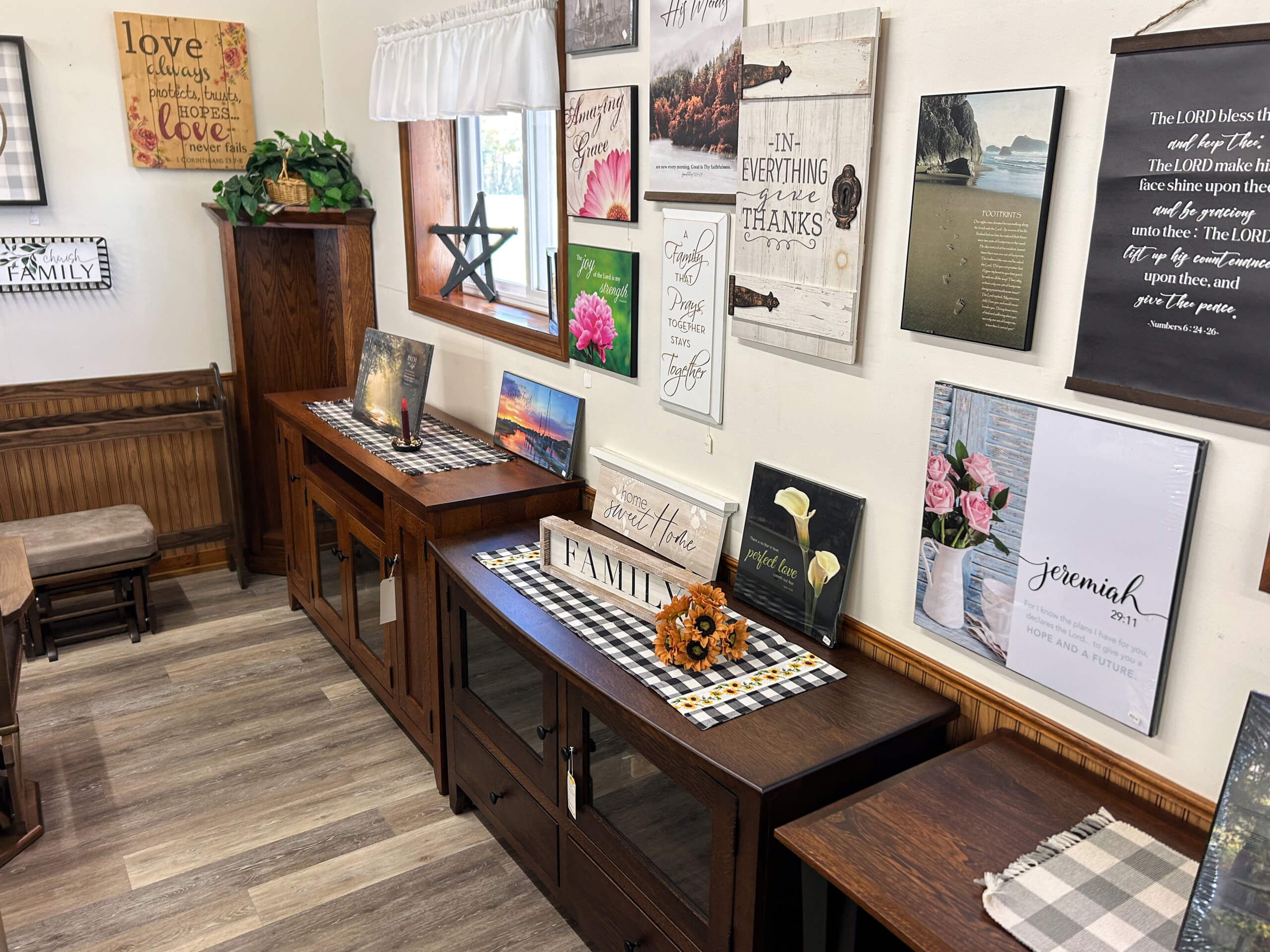 Yolder's Country Furniture Belle Center OH A room with wooden furniture, plaid table runners, and wall art featuring inspirational quotes and floral images, next to a window with white curtains.