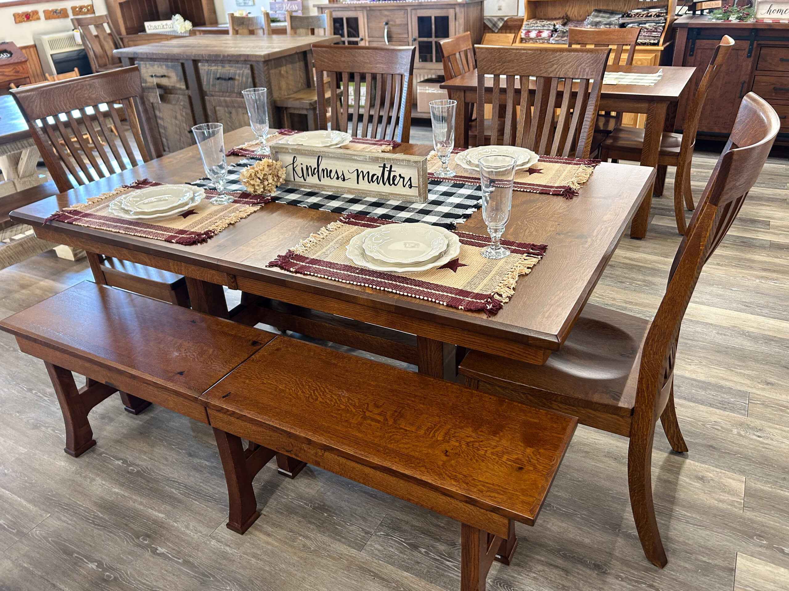 Yolder's Country Furniture Belle Center OH A wooden dining table set with four chairs and a bench, set for six with plates, glasses, placemats, and a centerpiece sign reading "Kindness matters.