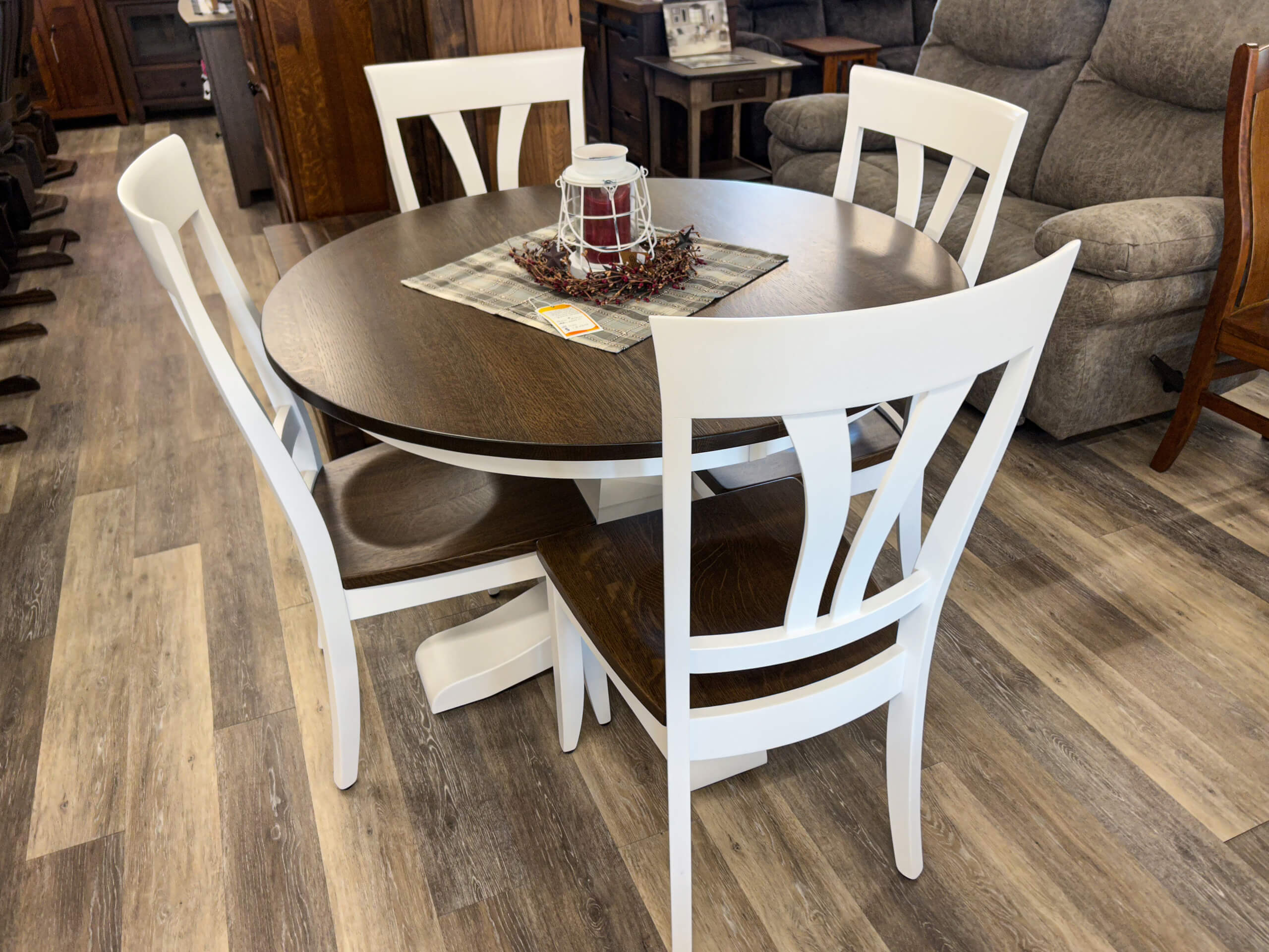 Yolder's Country Furniture Belle Center OH A round wooden table with a dark top and four white chairs, set on a wood-patterned floor with a centerpiece and placemat in a furniture store.