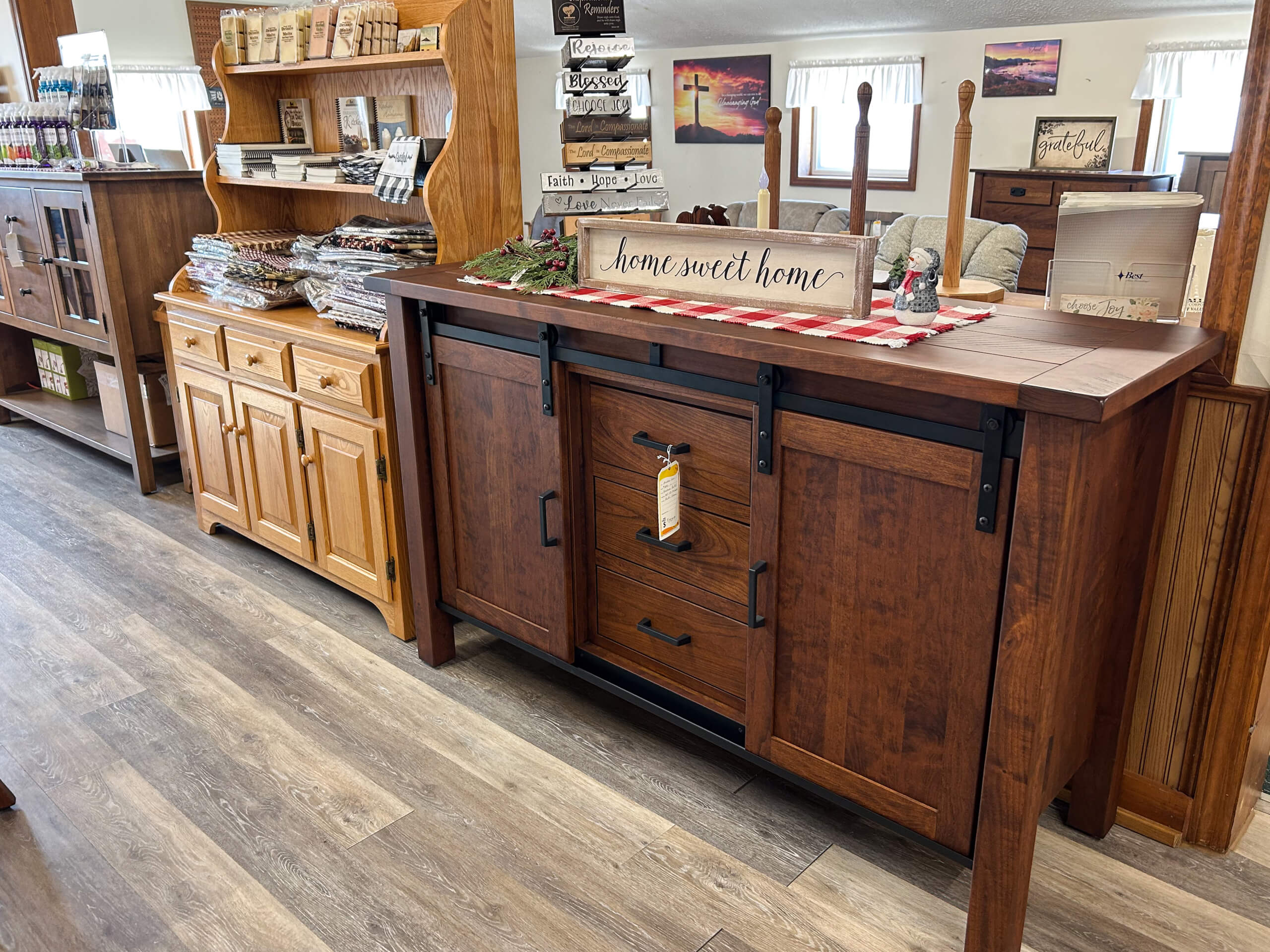 Yolder's Country Furniture Belle Center OH A wooden sideboard with sliding doors is displayed in a store, next to a hutch filled with packaged goods and decor items; a "Home Sweet Home" sign sits on top.