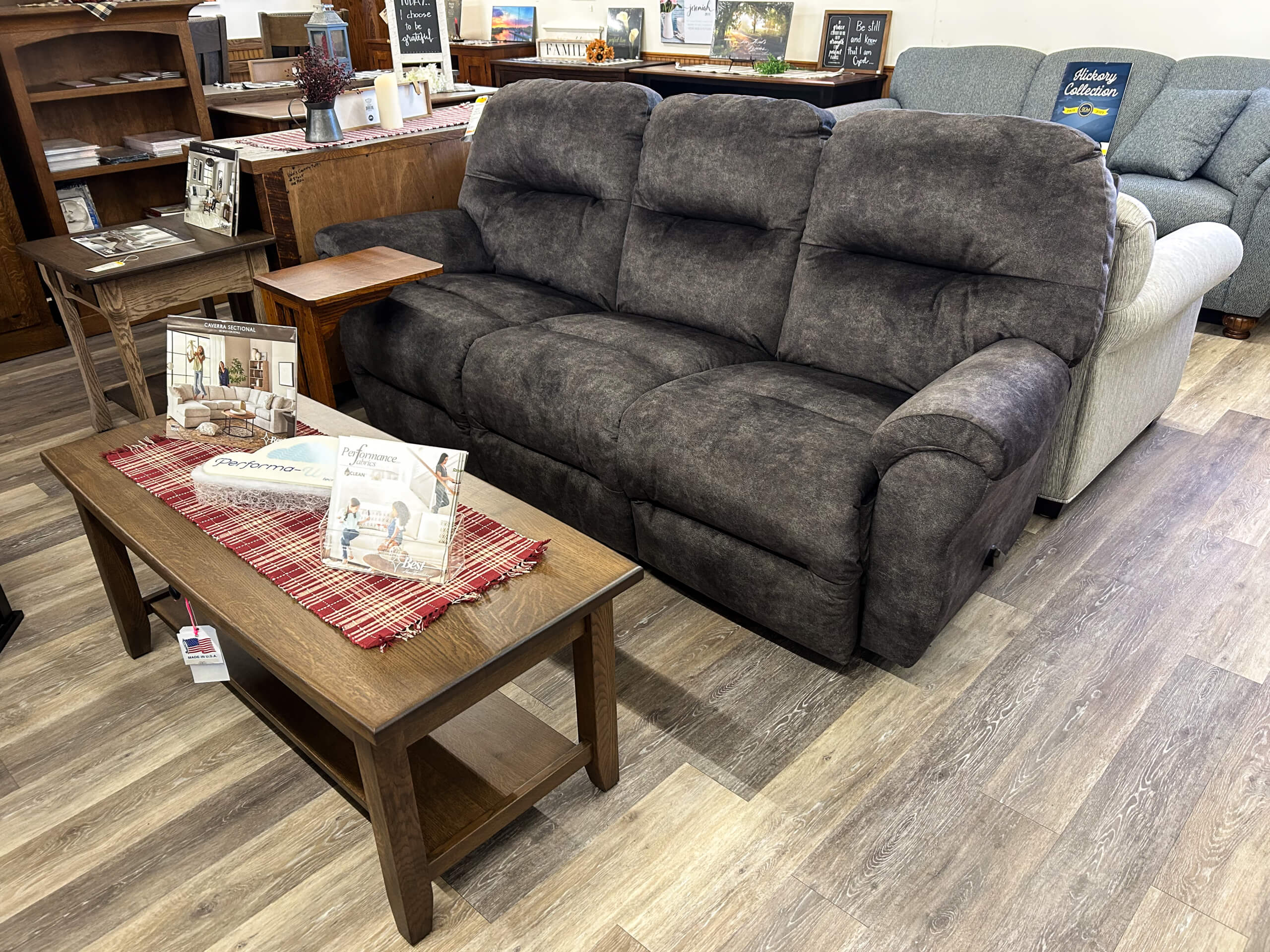 Yolder's Country Furniture Belle Center OH A dark gray upholstered reclining sofa is displayed in a showroom next to a wooden coffee table with brochures and a red plaid runner on top.