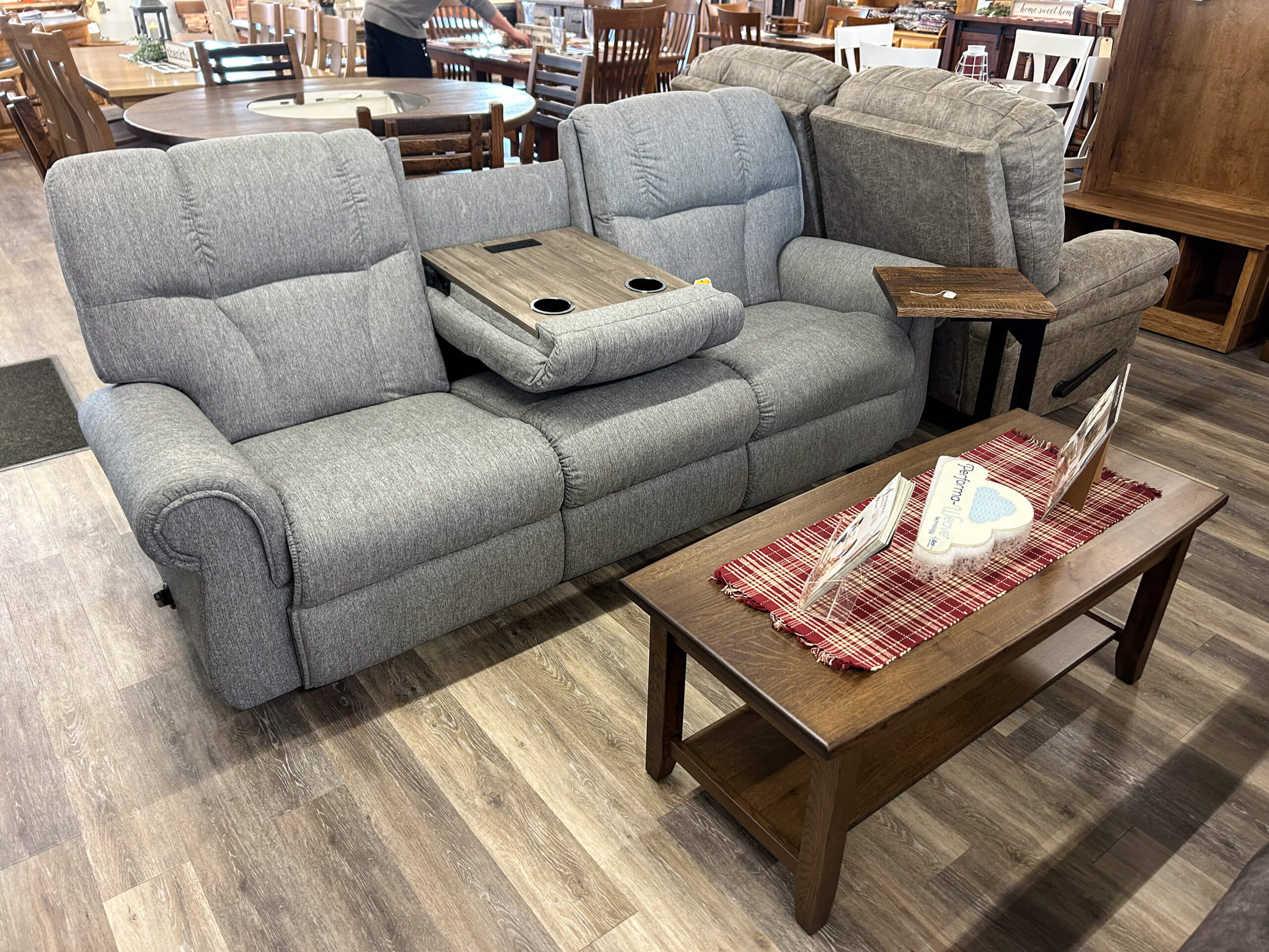 Yolder's Country Furniture Belle Center OH Gray upholstered reclining sofa with a center console and cup holders, positioned next to a wooden coffee table with a red plaid runner and decorative items in a showroom.