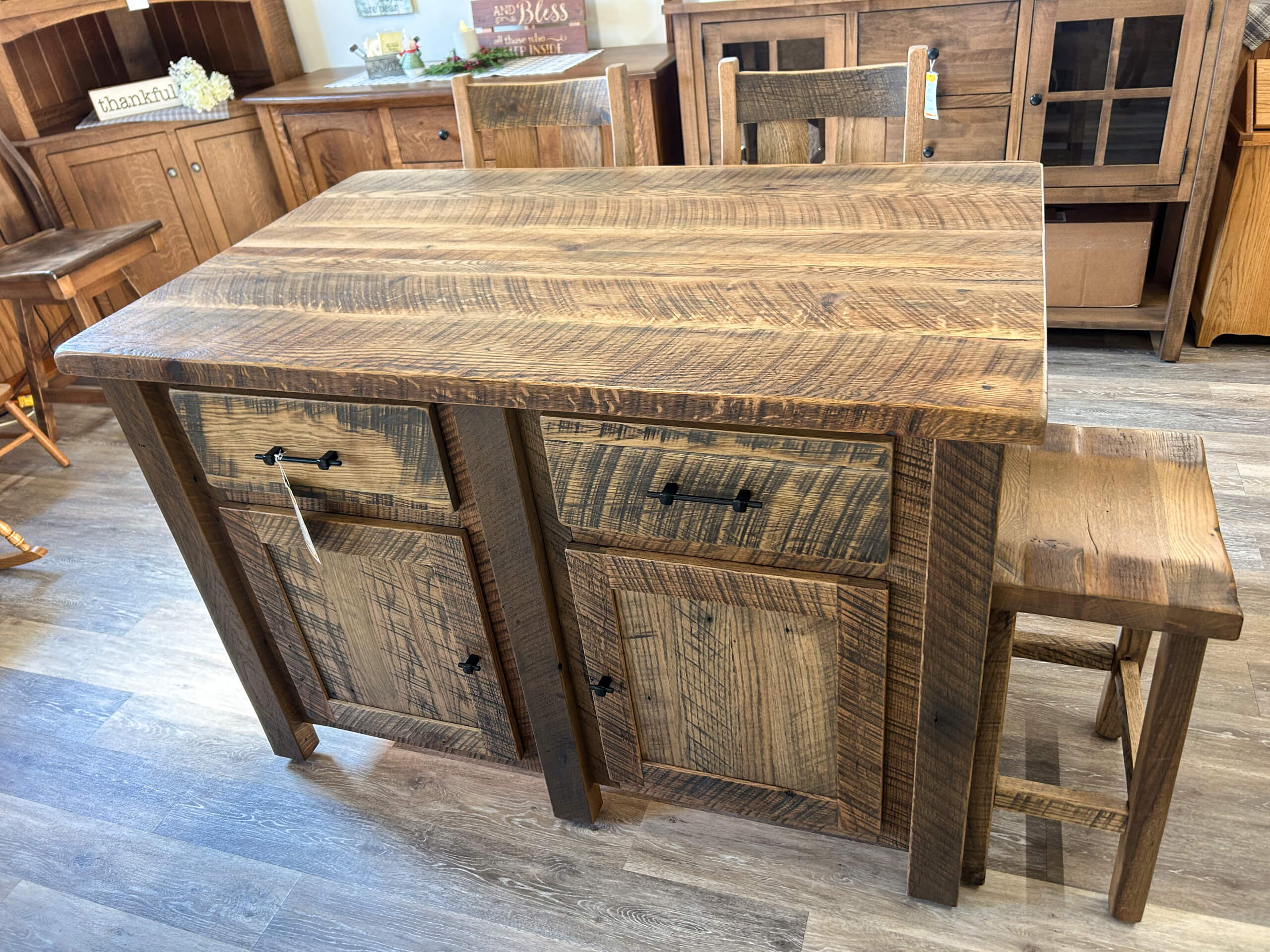 Yolder's Country Furniture Belle Center OH A rustic wooden kitchen island with two drawers, two cabinets, and a matching stool, placed on a wooden floor in a furniture showroom.