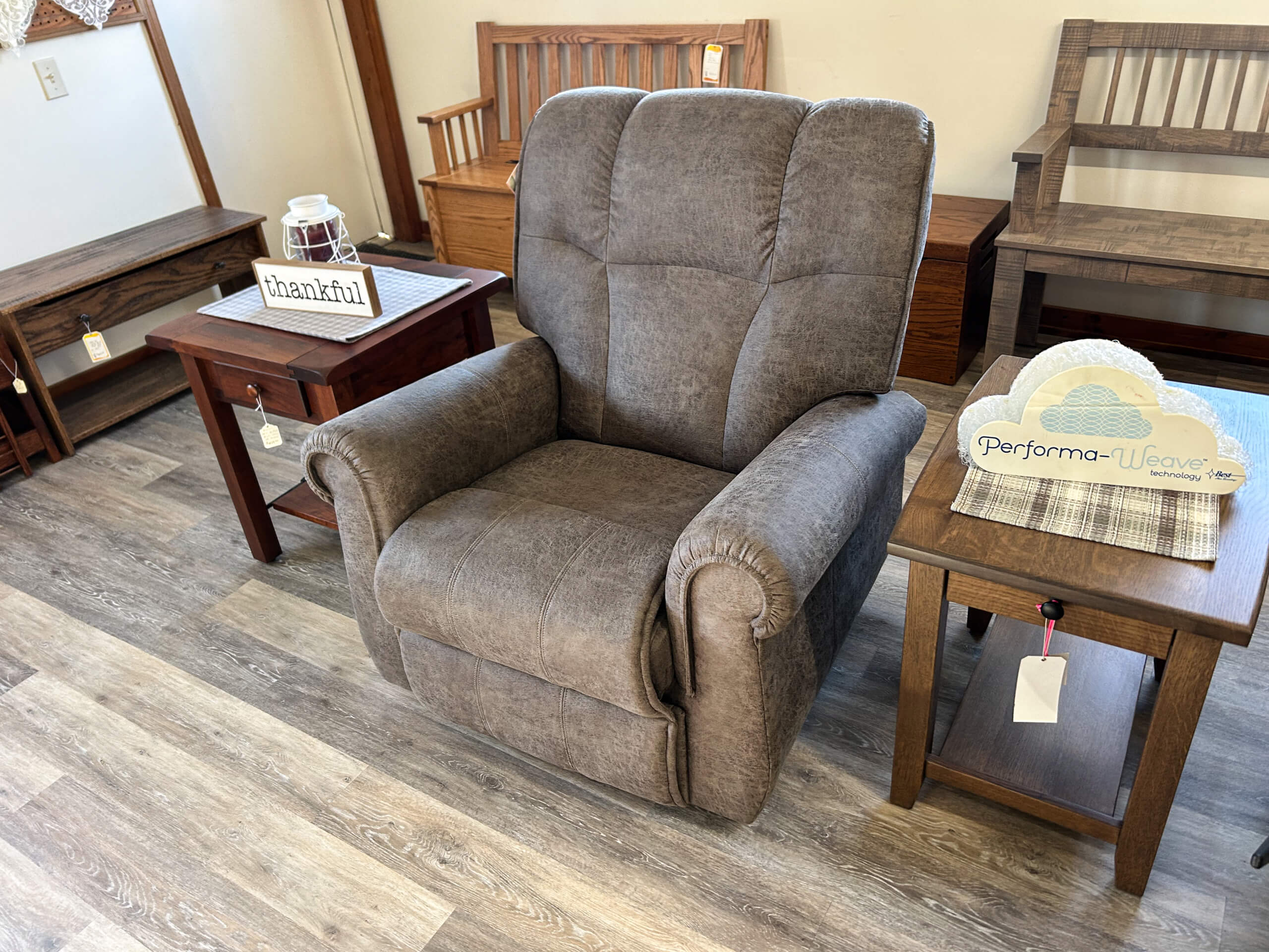 Yolder's Country Furniture Belle Center OH A gray upholstered recliner is displayed between two wooden side tables in a furniture showroom with a wood-look floor.