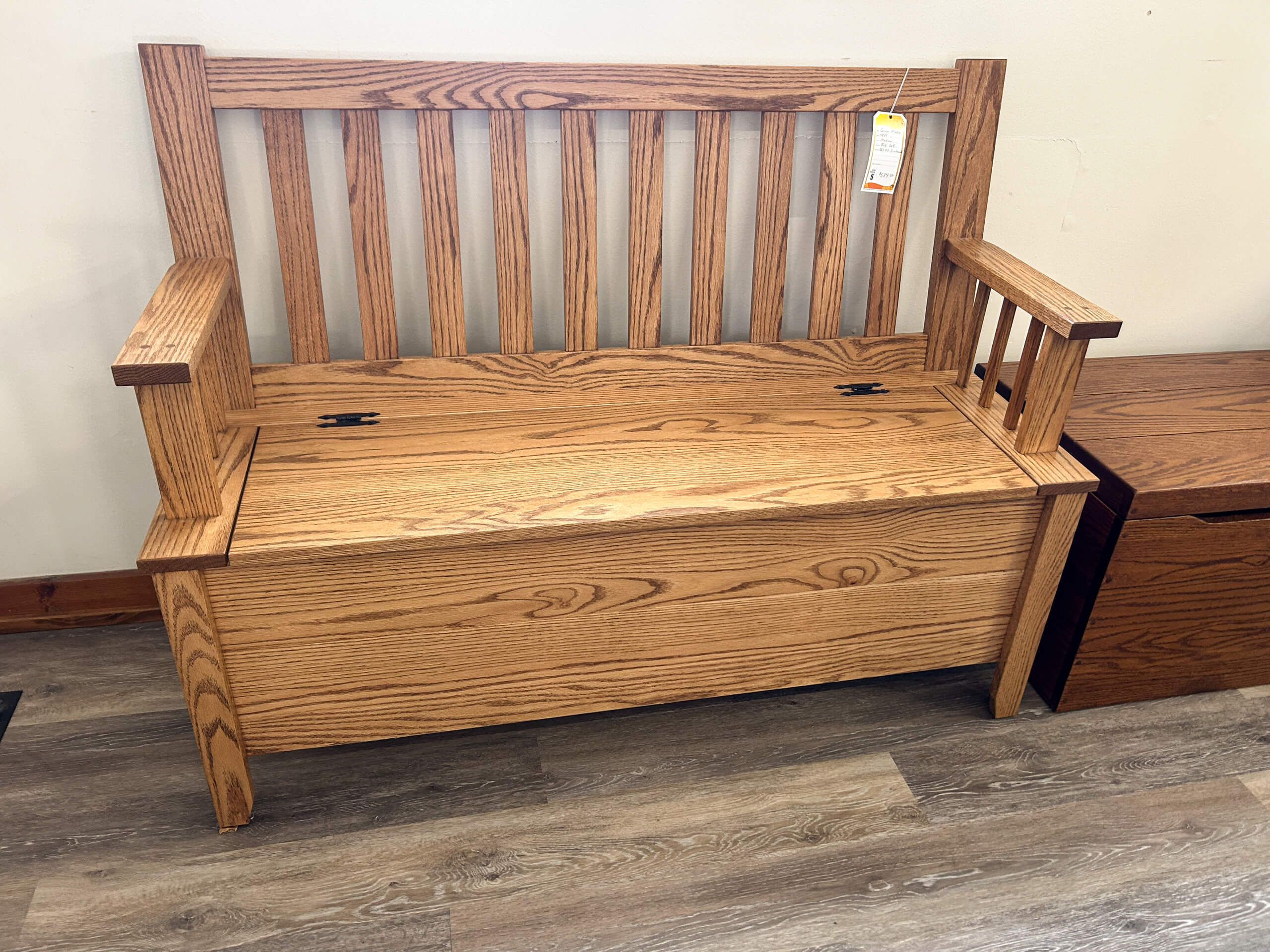 Yolder's Country Furniture Belle Center OH Wooden bench with vertical slats on the backrest, armrests, and a hinged storage compartment under the seat, placed on a wood-look floor next to a wooden cabinet.
