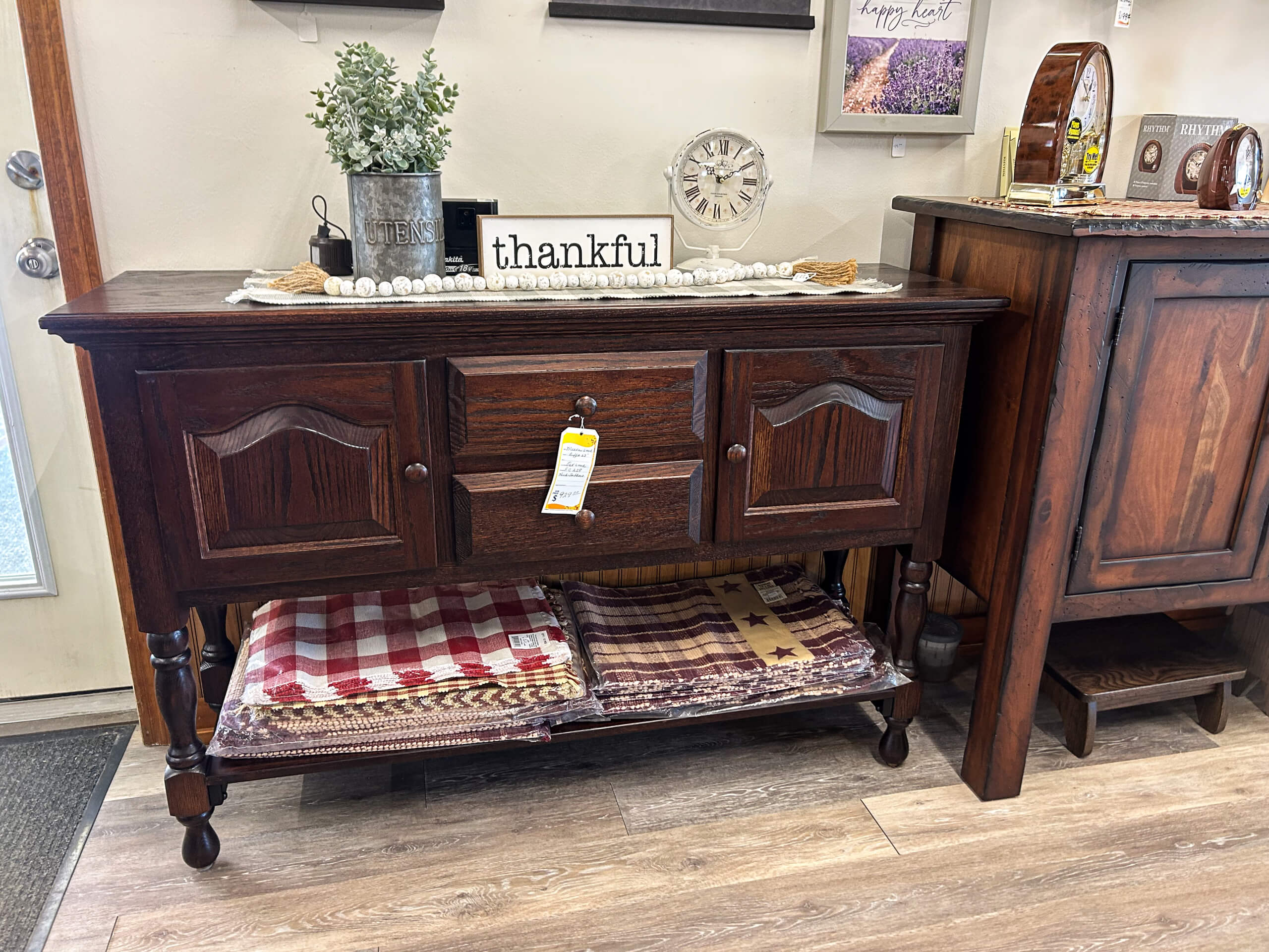 Yolder's Country Furniture Belle Center OH A dark wood sideboard with two cabinets and a drawer, decorated with a plant, clock, and "thankful" sign, with folded quilts on a lower shelf.
