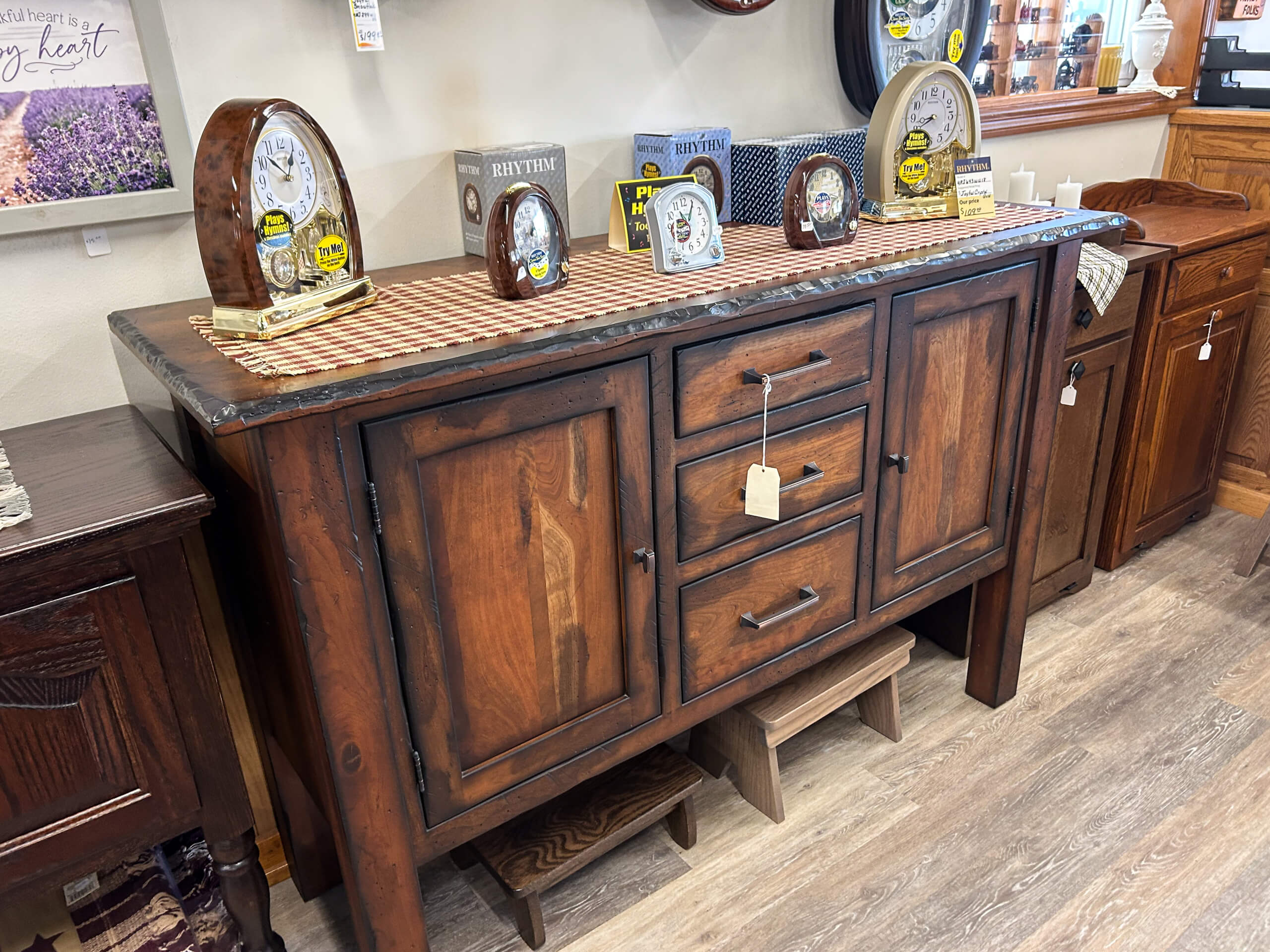 Yolder's Country Furniture Belle Center OH A wooden sideboard with three drawers and two cabinets is displayed in a store, topped with decorative clocks and a patterned table runner.