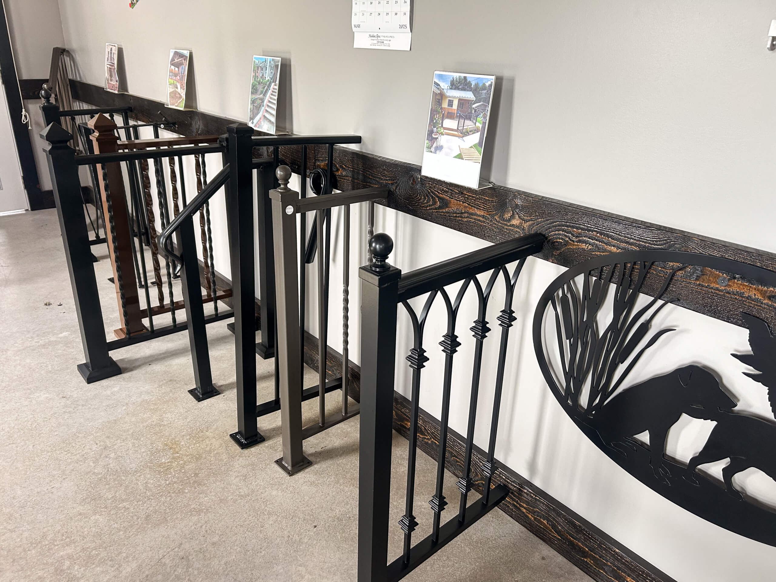 Zook's Metal Fabricating & Railings Allenwood, PA Several styles of metal railings are displayed upright against a wall, each with unique designs. Framed photos and a calendar hang above the railings.