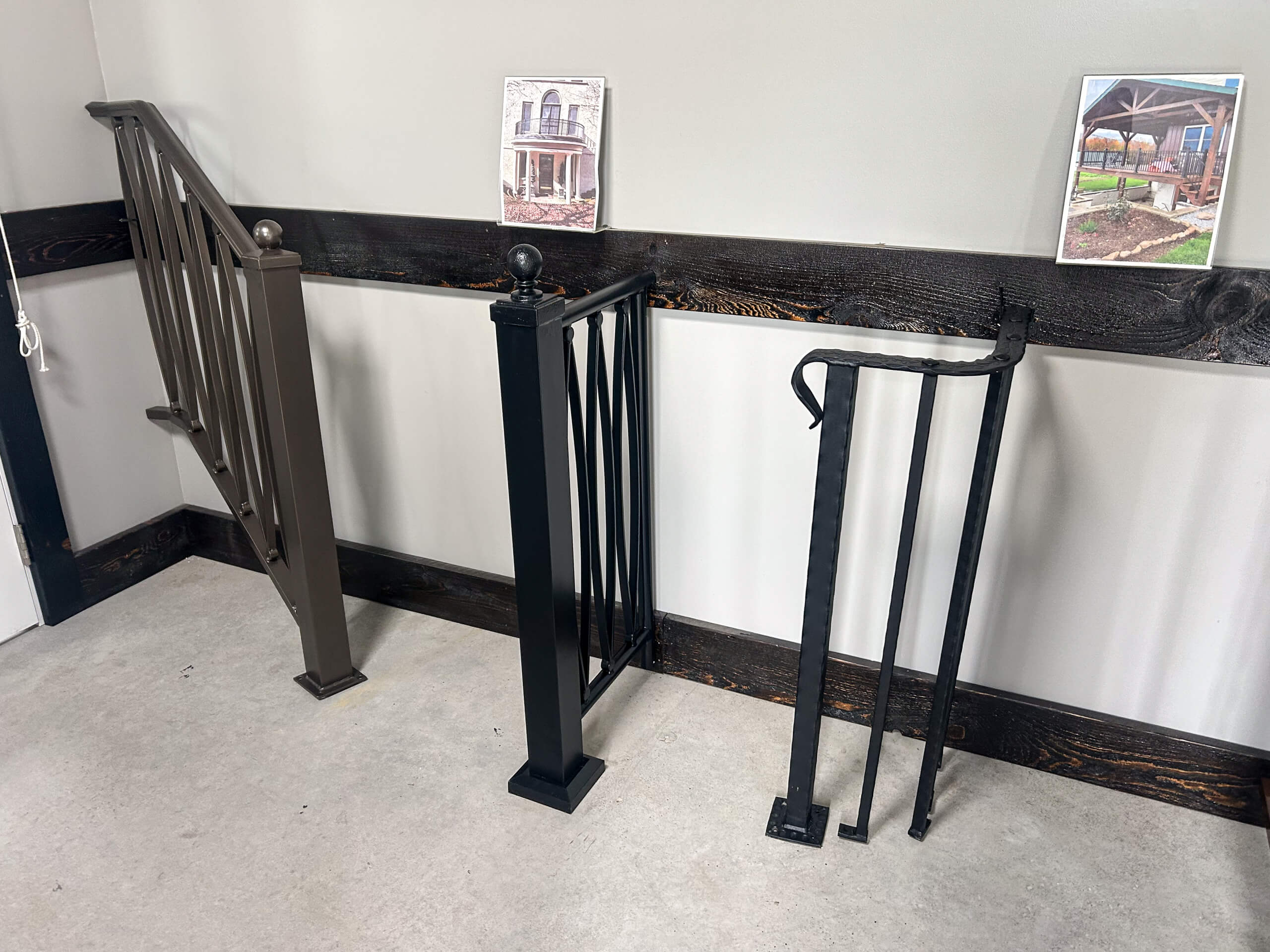 Zook's Metal Fabricating & Railings Allenwood, PA Three different metal railing samples are displayed upright against a wall, with two framed photos mounted above on a wooden trim.