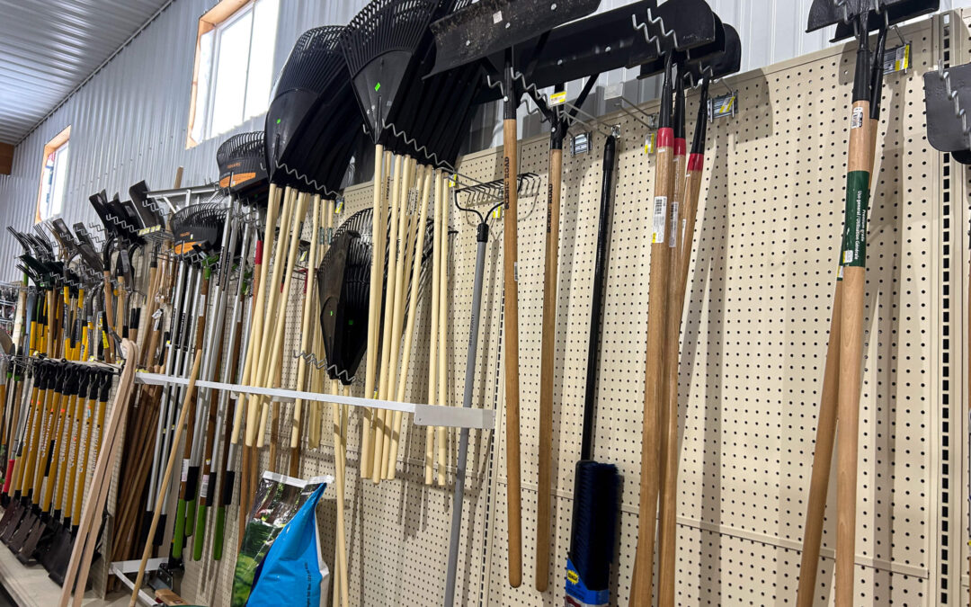 Shetler's Supplies Hardware Variety Bulk Foods Greenville PA Garden rakes and other long-handled tools are displayed on a pegboard wall in a store aisle, with bags of soil or mulch on the shelf below.