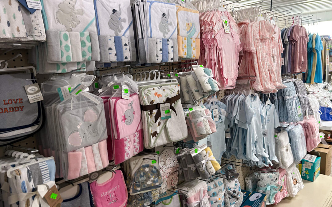 Shetler's Supplies Hardware Variety Bulk Foods Greenville PA Shelves display assorted baby clothes, bibs, and blankets in pastel colors, including pink, blue, and gray, with animal prints and text designs.
