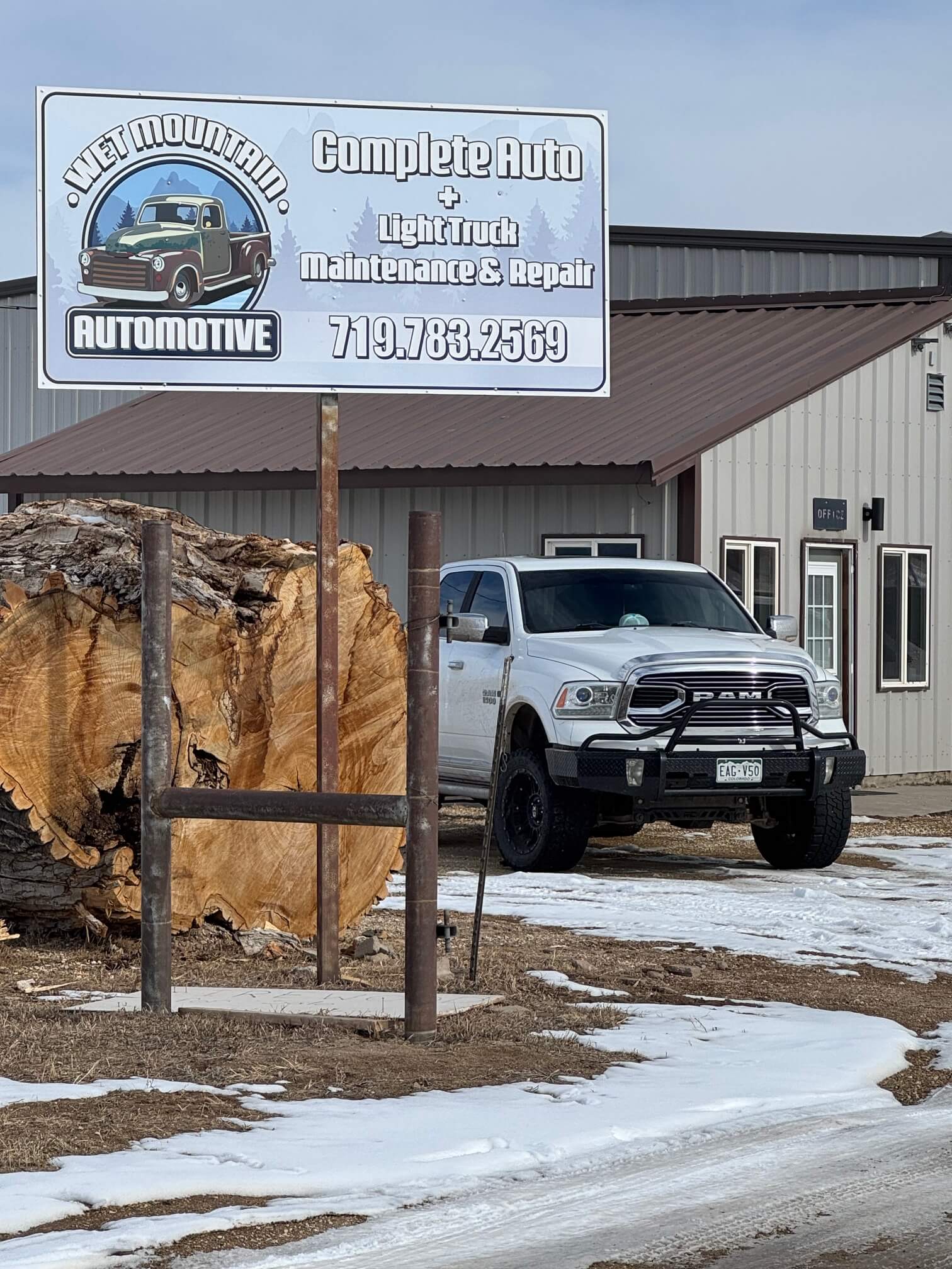 A white pickup truck is parked outside a metal building near a large log and a sign for "Wet Mountain Automotive" advertising auto maintenance and repair services.