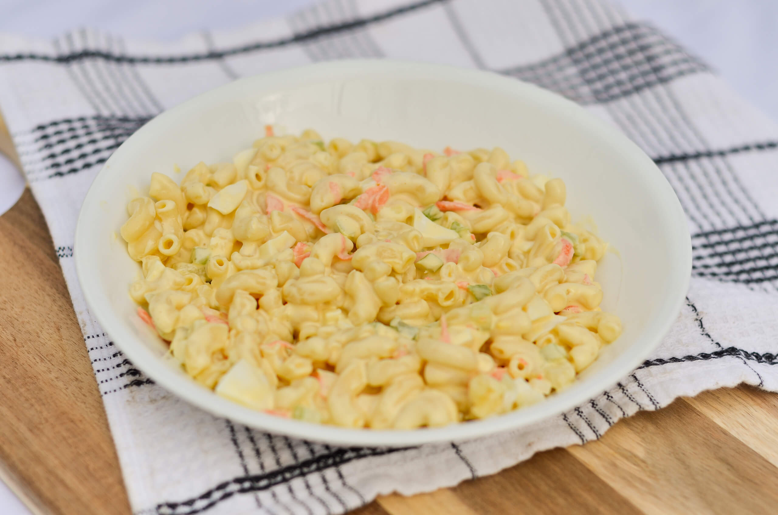 Nolt's Country Market Eldon, Missouri A white bowl filled with macaroni salad containing elbow pasta and chopped vegetables sits on a checked towel over a wooden surface.