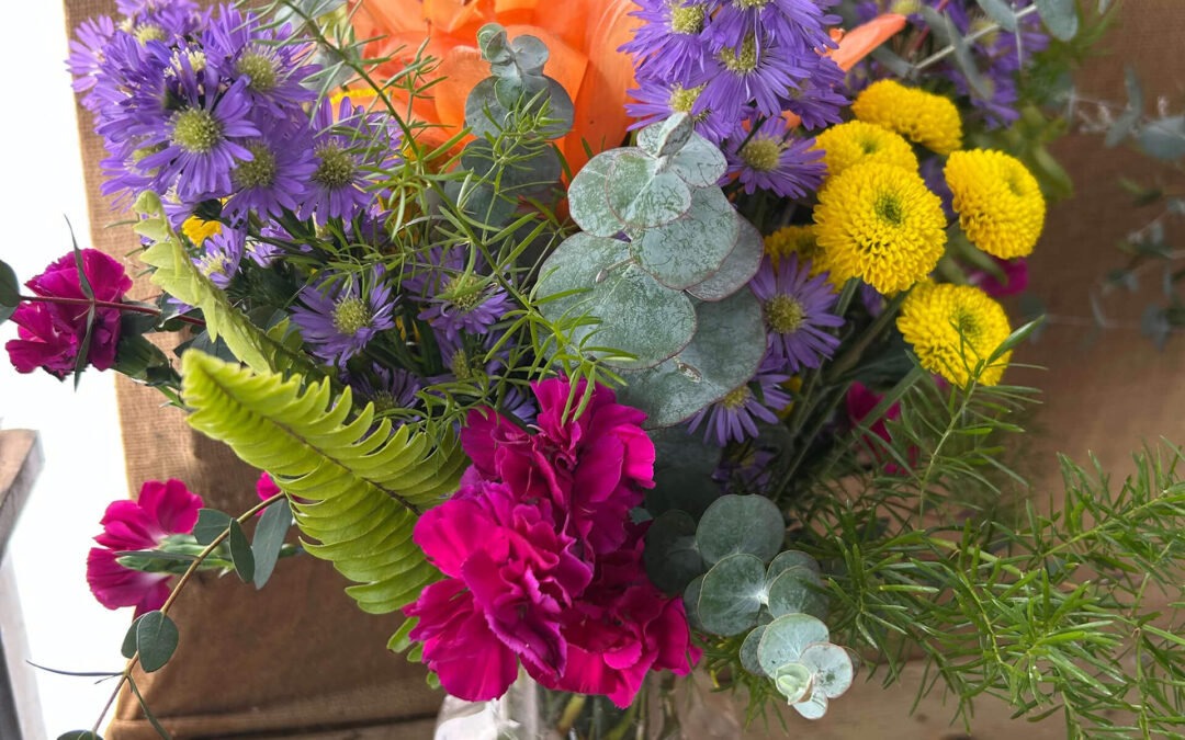 2.11.26 Swampy Hollow Greenhouse Valentines Gallery 1 A clear glass vase holds a mixed bouquet of purple, yellow, pink, and orange flowers with assorted greenery, set on a wooden surface against a burlap background.
