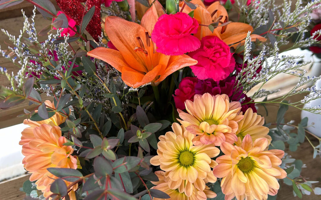 2.11.26 Swampy Hollow Greenhouse Valentines Gallery 2 A floral arrangement with orange lilies, yellow daisies, pink carnations, red heart decorations, and assorted greenery on a wooden surface.