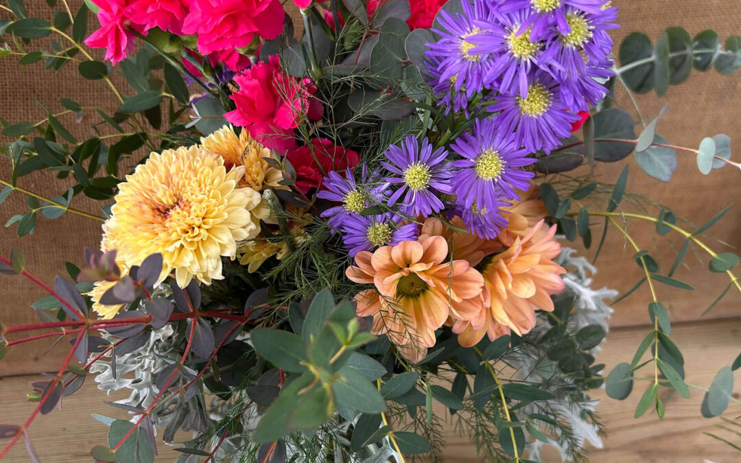 2.11.26 Swampy Hollow Greenhouse Valentines Gallery 3 A bouquet of mixed flowers including purple, red, yellow, and orange blooms with greenery, placed on a wooden surface. A "Happy Valentine's Day" sign is in the arrangement.
