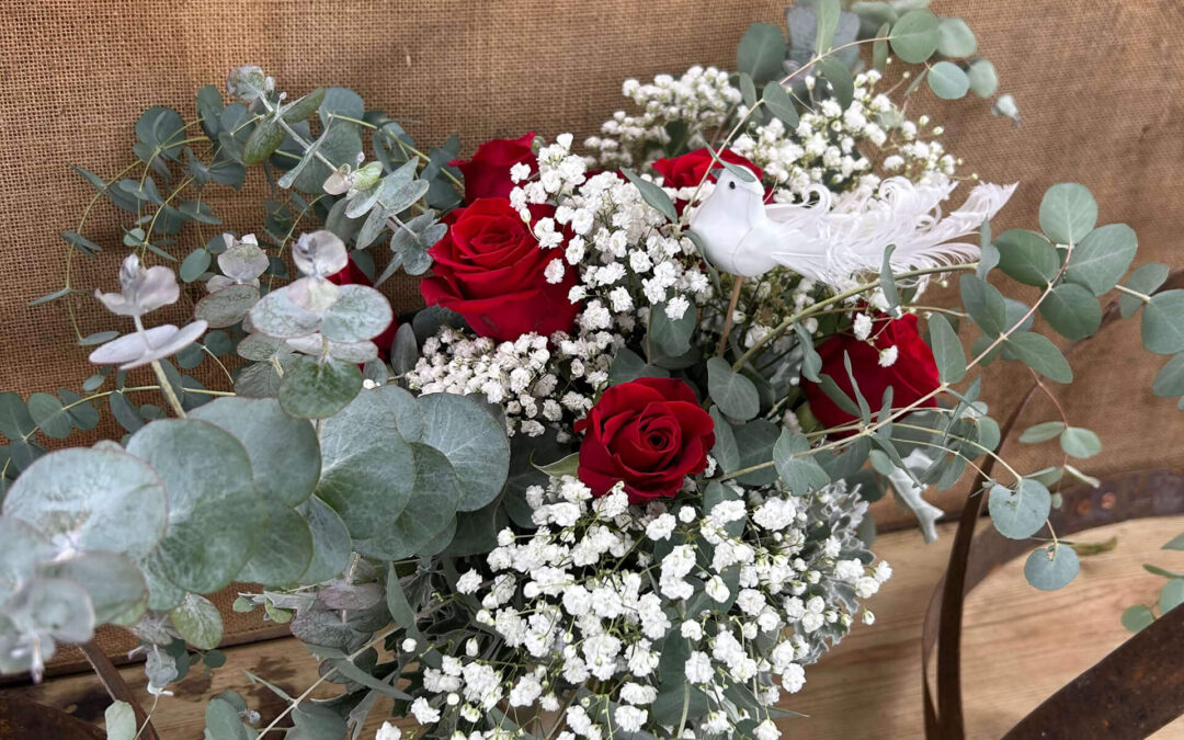 2.11.26 Swampy Hollow Greenhouse Valentines Gallery 4 A floral arrangement with red roses, baby's breath, eucalyptus leaves, and a white decorative bird, set against a burlap background.