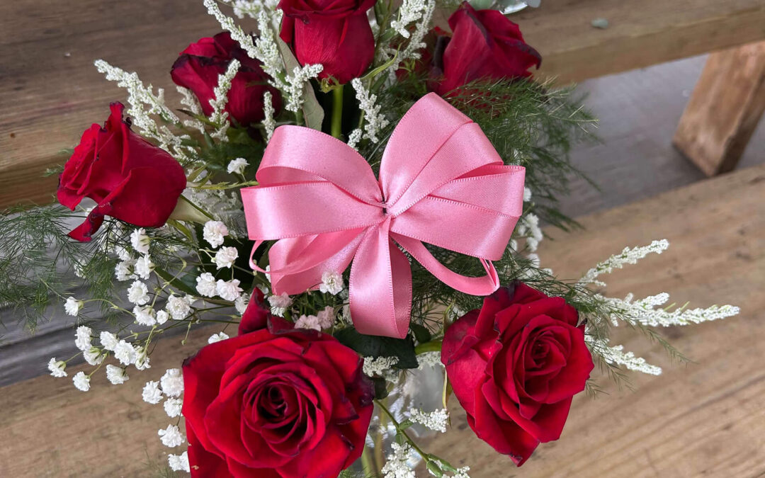 2.11.26 Swampy Hollow Greenhouse Valentines Gallery 7 A bouquet of six red roses, white filler flowers, and greenery is arranged with a large pink ribbon bow, placed on wooden steps.