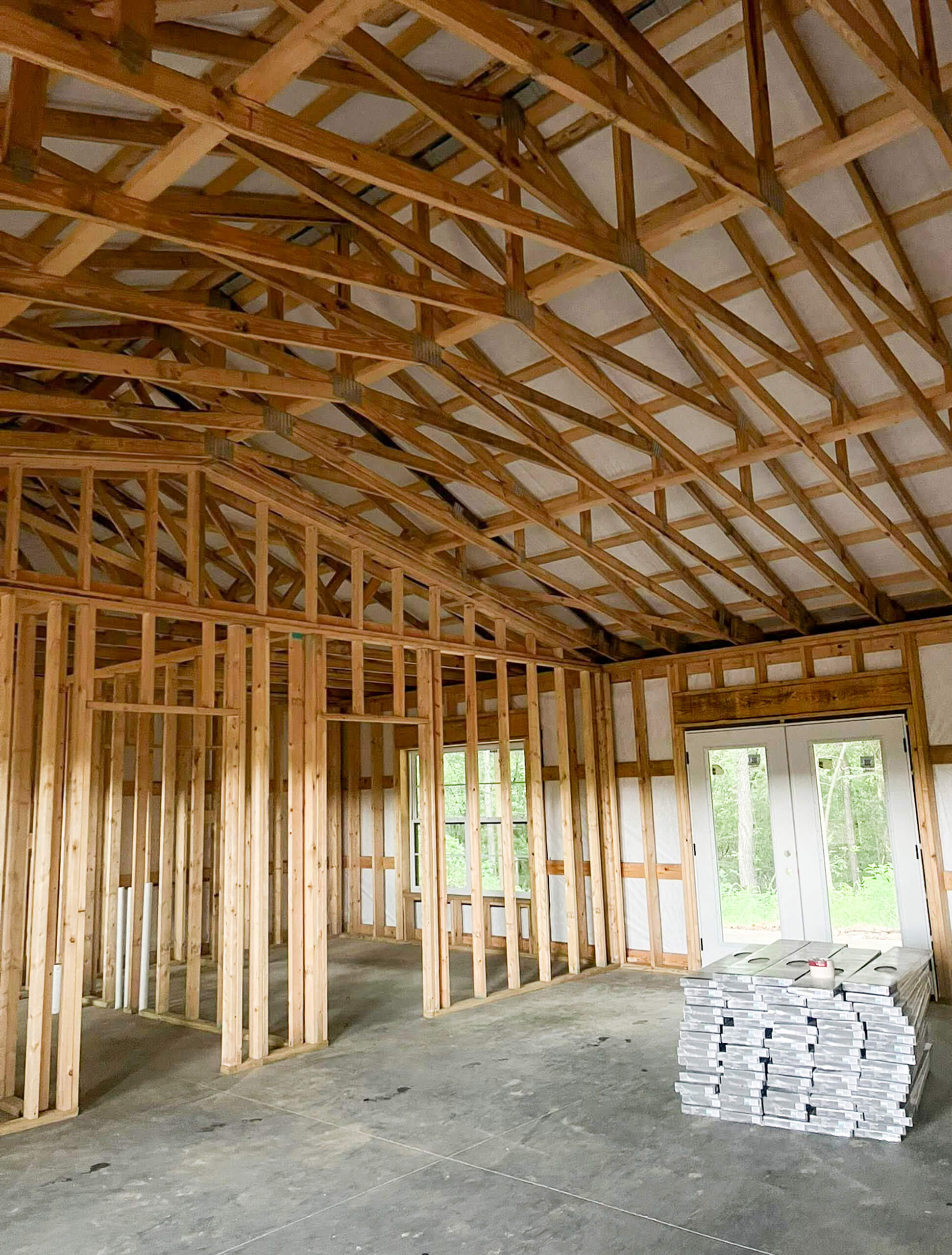 Bud's Construction Miunfordville KY Interior view of a house under construction with exposed wooden framing, unfinished walls, and a stack of drywall sheets on the concrete floor. Double doors lead outside.