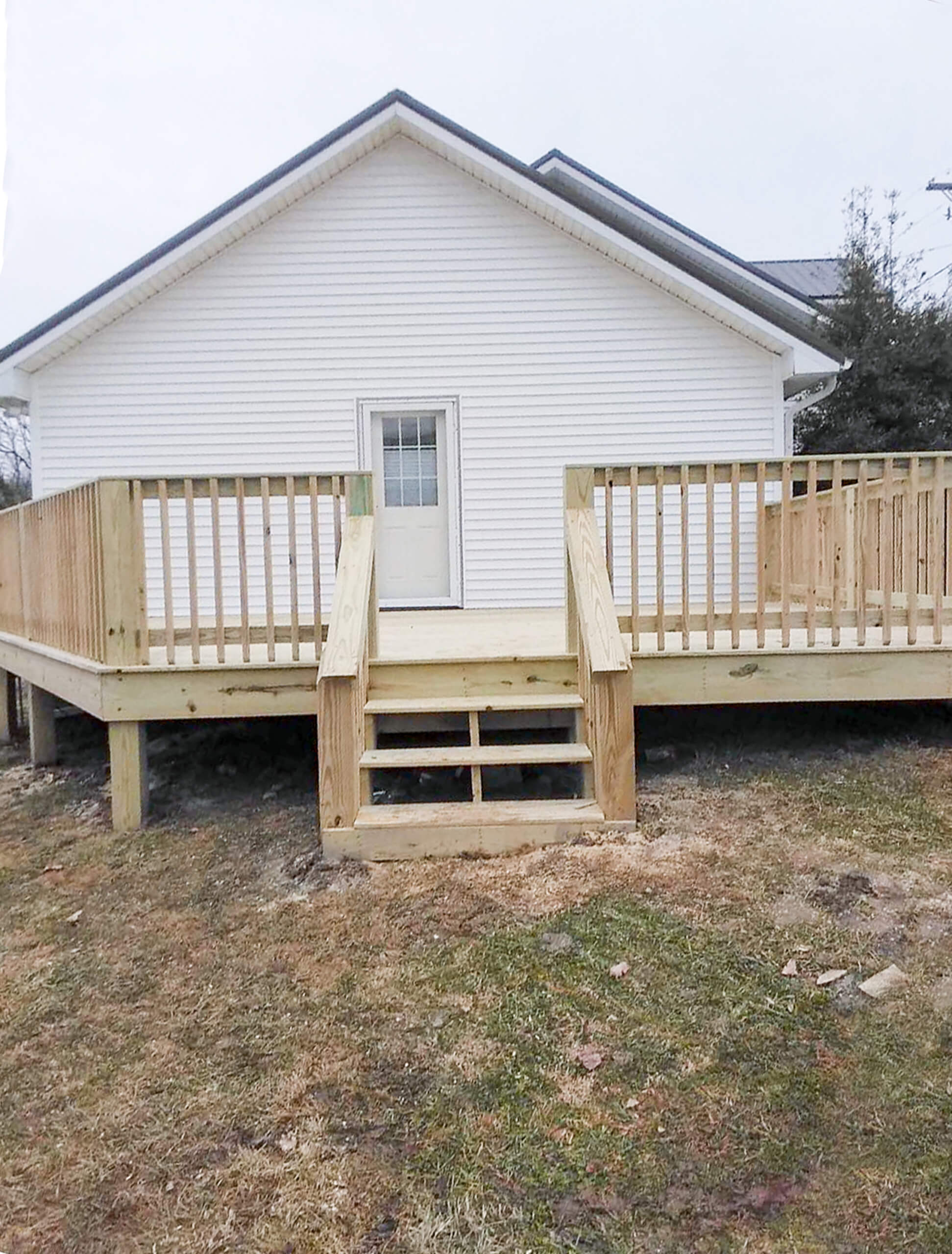 Bud's Construction Miunfordville KY A small wooden deck with railings and steps leads to a door at the back of a white house. The yard around the deck has patchy grass and bare soil.
