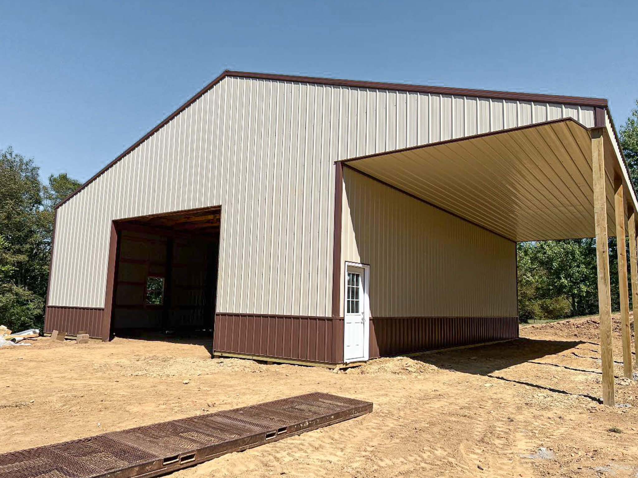 Bud's Construction Miunfordville KY A large metal building with a high roof, open bays, a white entry door, and brown trim, situated on a dirt lot with some trees in the background.