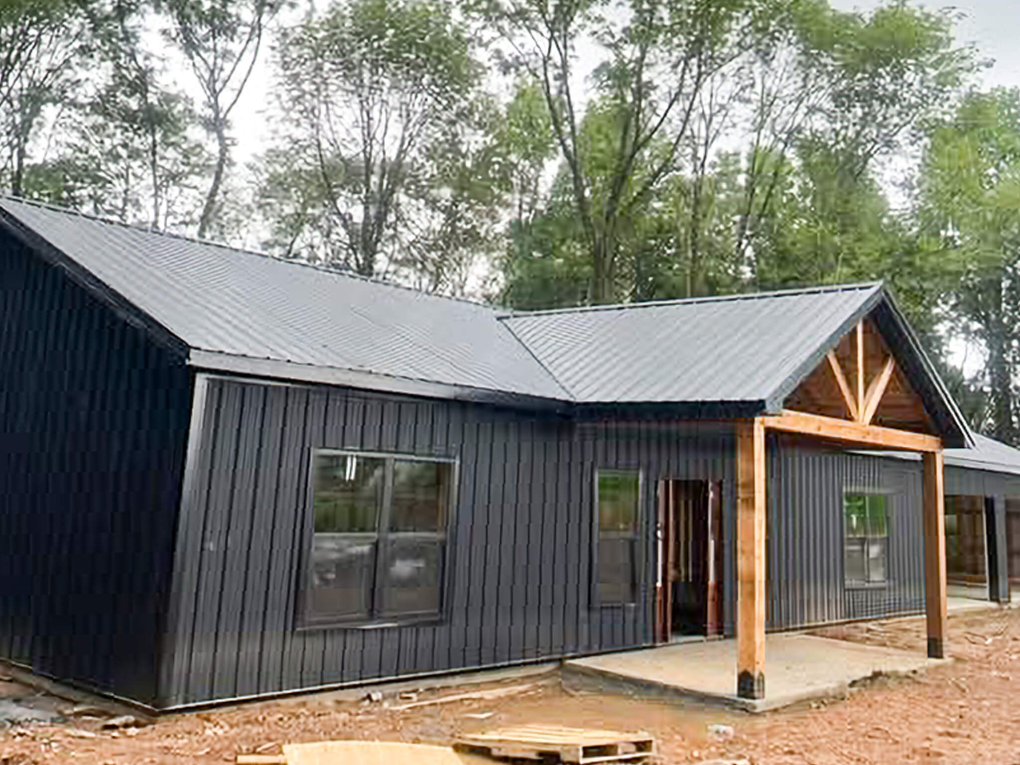 Bud's Construction Miunfordville KY A single-story modern house under construction with dark vertical siding, a metal roof, large windows, and a wooden porch frame, surrounded by trees.