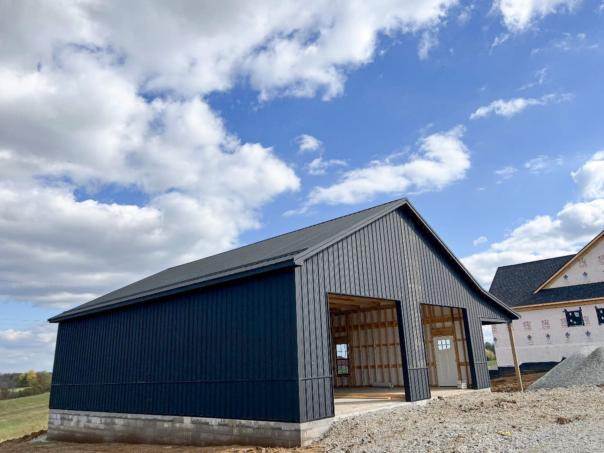 Bud's Construction Miunfordville KY A large, unfinished black metal building with open garage doors stands next to a partially constructed house under a partly cloudy sky.