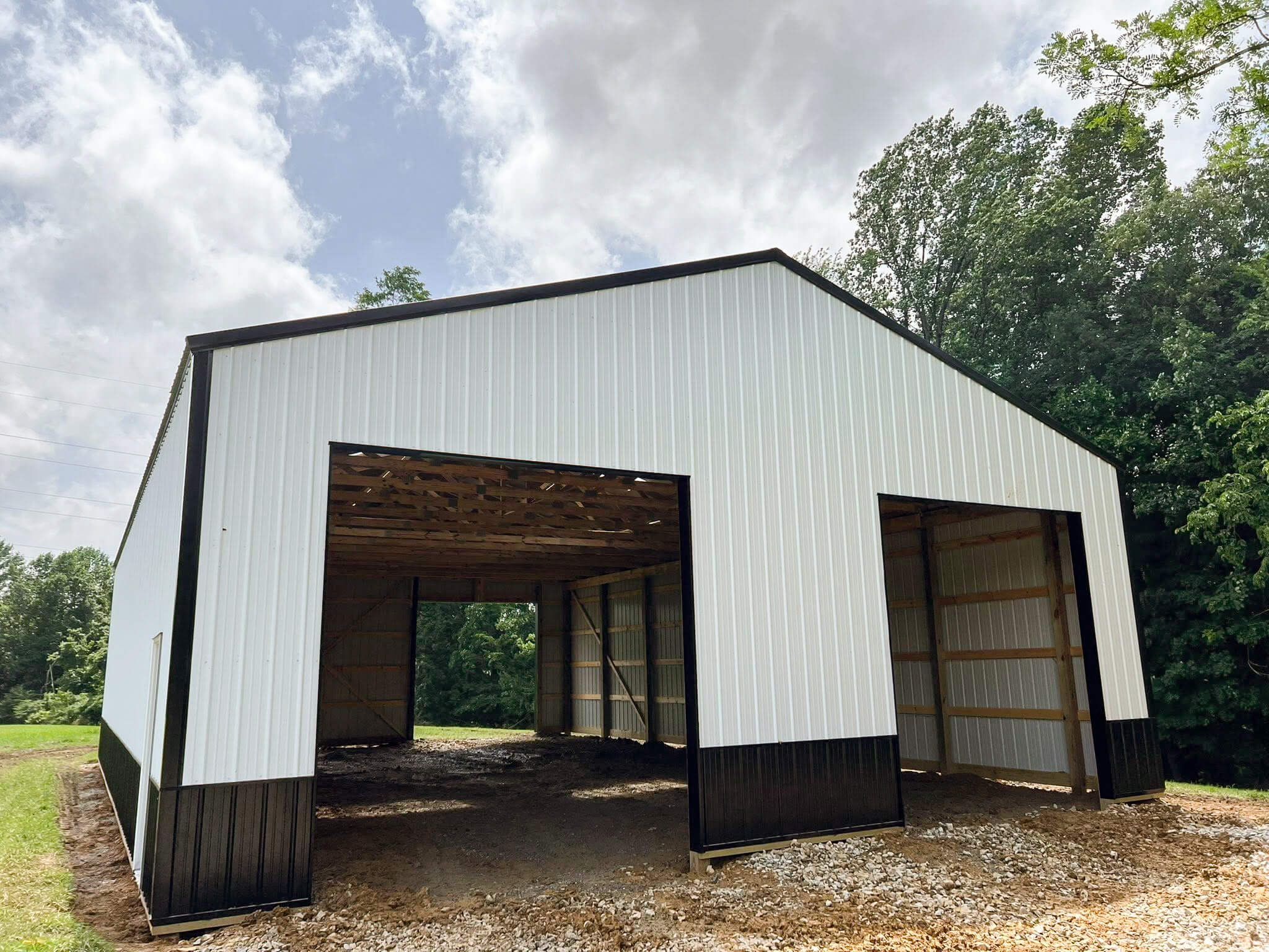 Bud's Construction Miunfordville KY A large metal building with two open garage doors, unfinished interior, white siding, and black trim, standing on a gravel foundation beside trees.