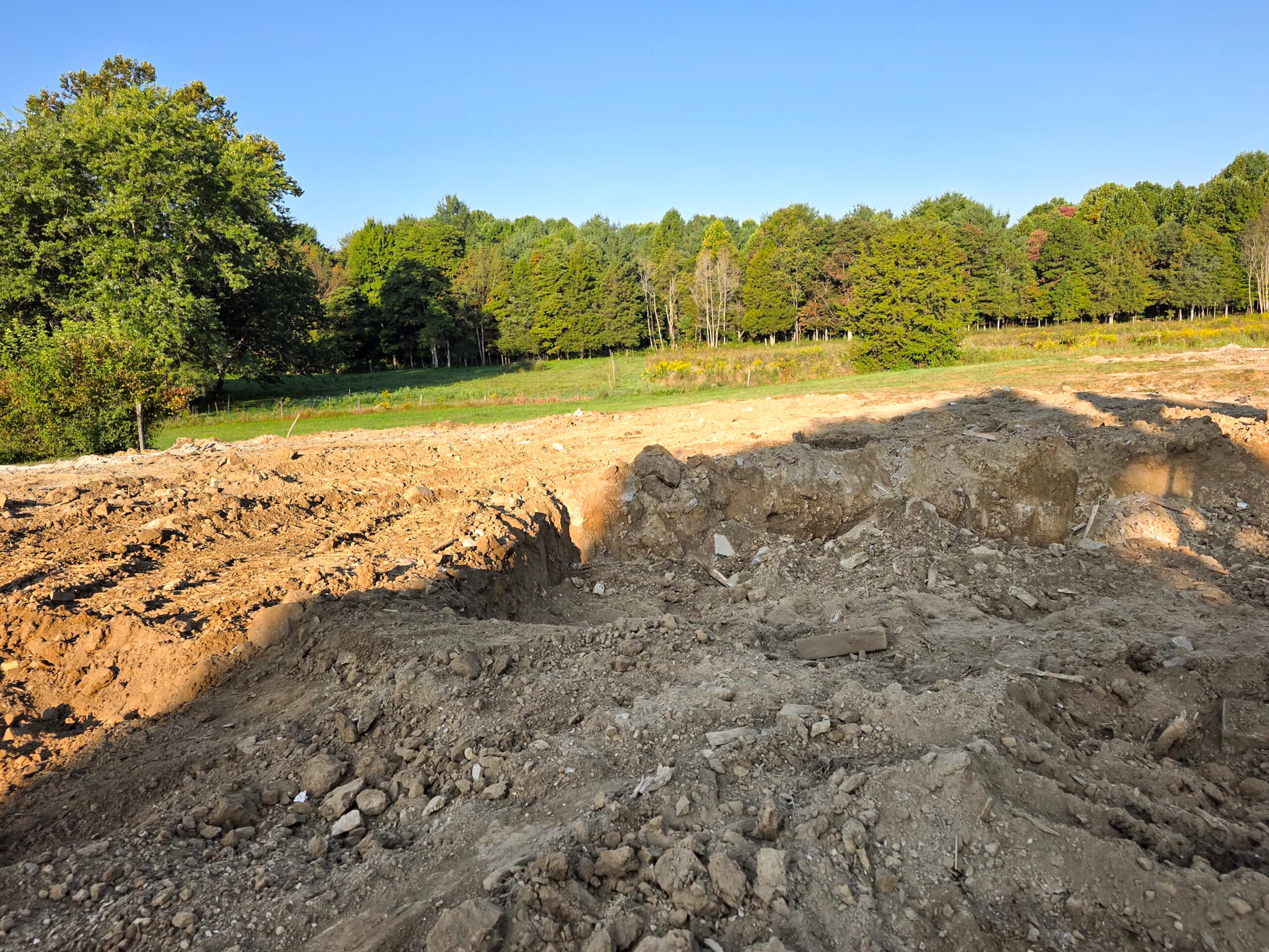 Elite Dumpster and Demolition Odon, IN A large dirt excavation site is in the foreground with grassy field and dense trees in the background under a clear blue sky.
